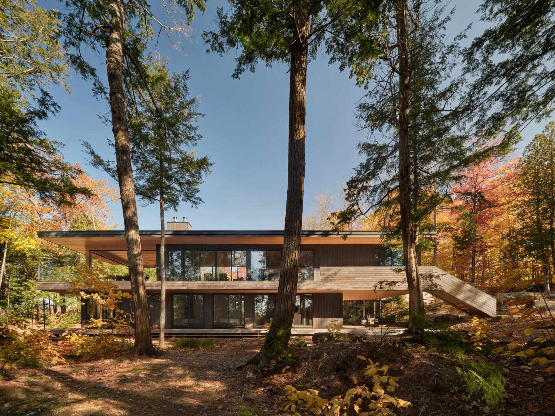 An off-grid cottage set on a forested peninsula, where a bridge leads to an elevated living level that sits lightly among the trees. Clad in greyed cedar and charcoal-stained timber, the exterior blends into the landscape while expansive glazing opens the home to water views on three sides.