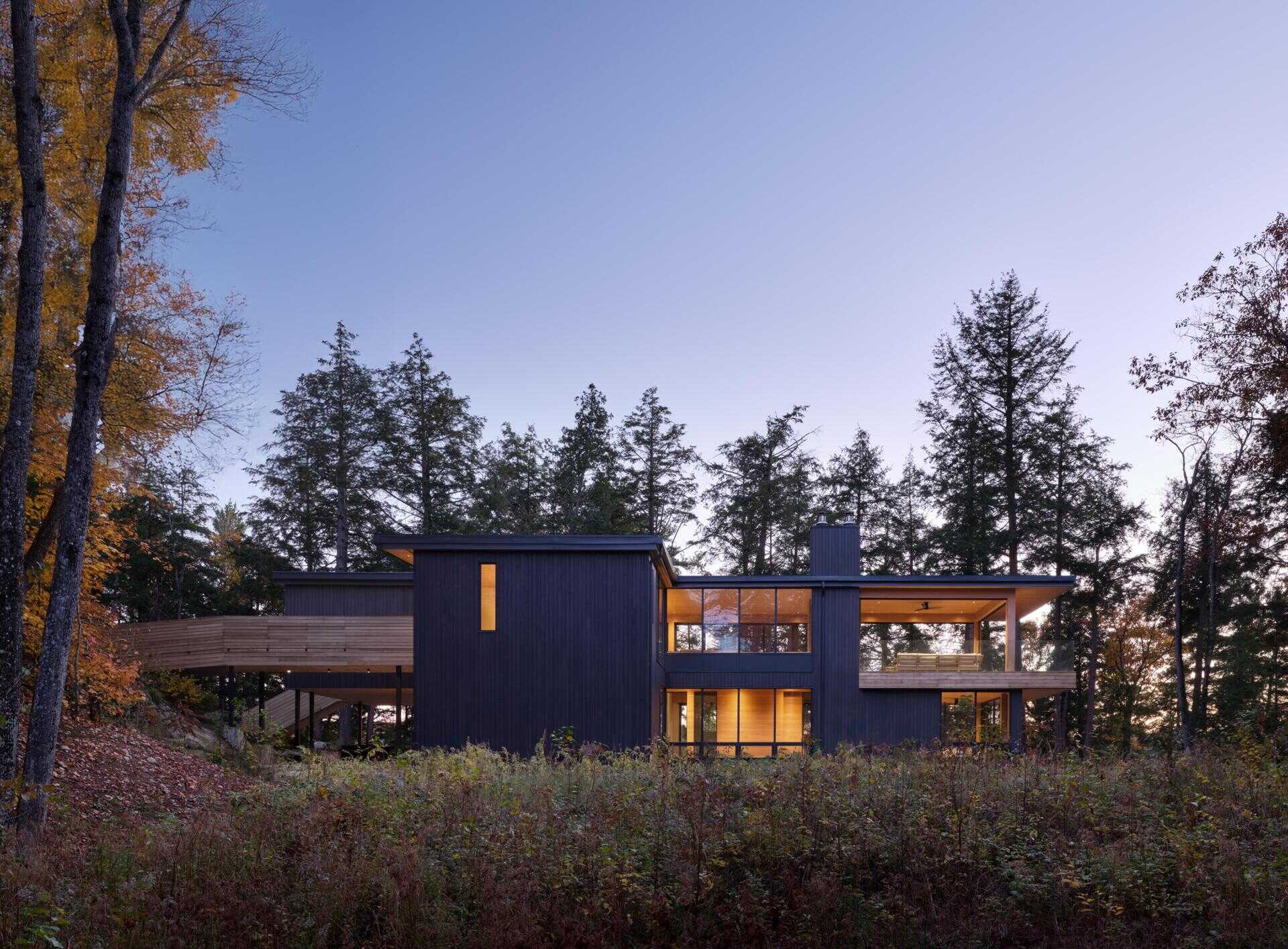 An off-grid cottage set on a forested peninsula, where a bridge leads to an elevated living level that sits lightly among the trees. Clad in greyed cedar and charcoal-stained timber, the exterior blends into the landscape while expansive glazing opens the home to water views on three sides.