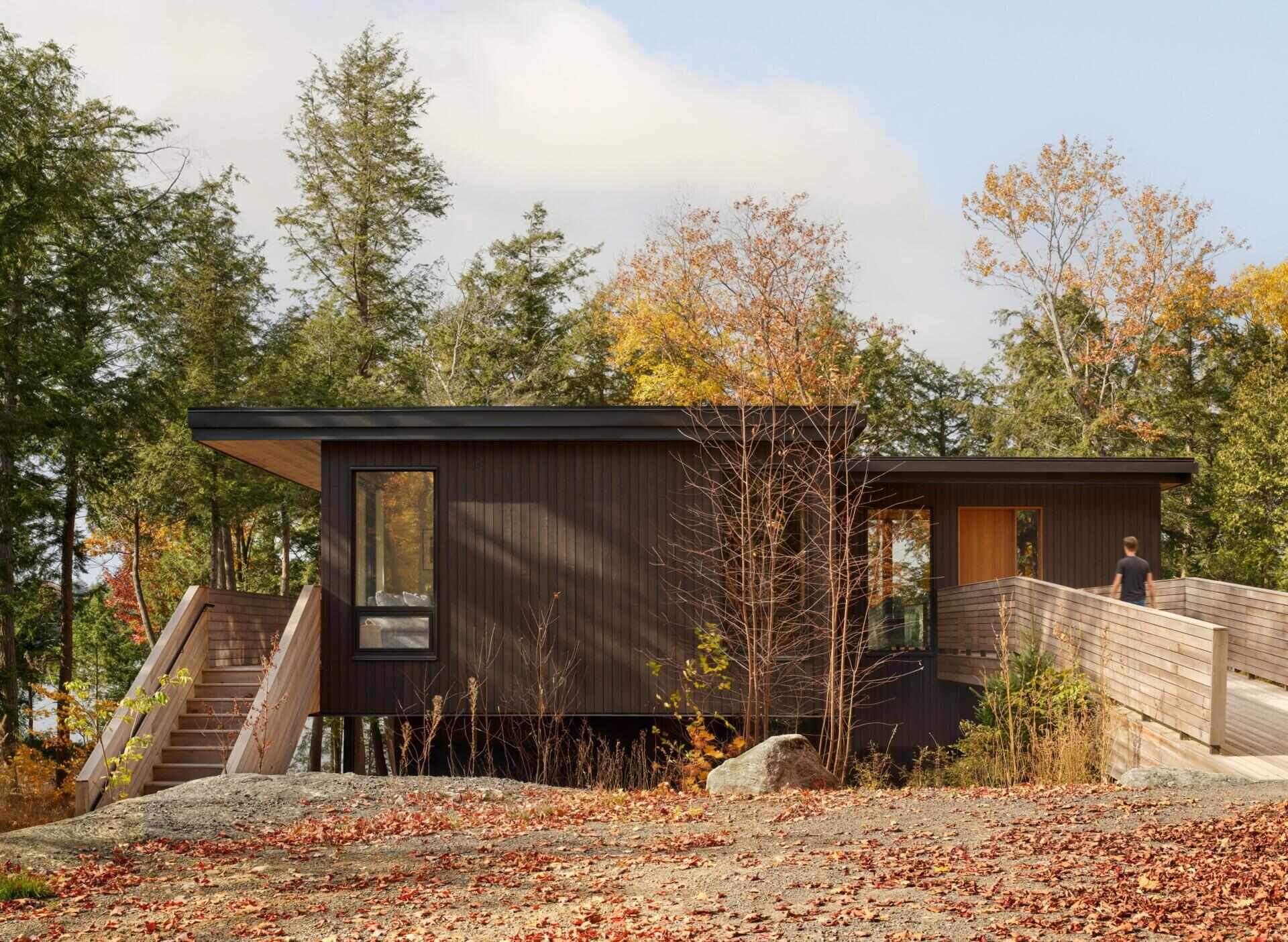 An off-grid cottage set on a forested peninsula, where a bridge leads to an elevated living level that sits lightly among the trees. Clad in greyed cedar and charcoal-stained timber, the exterior blends into the landscape while expansive glazing opens the home to water views on three sides.