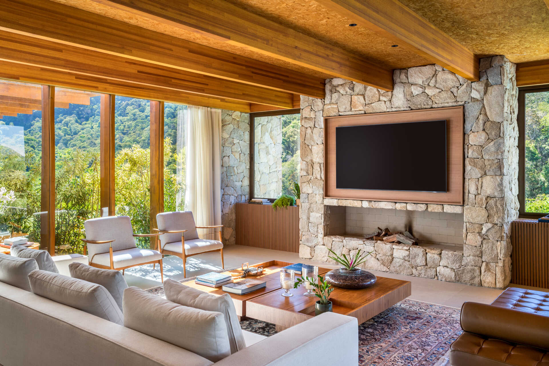 This living room blends warm wood and rugged stone, while floor-to-ceiling windows bring in mountain views and daylight, softened by airy curtains. Low wood tables and a patterned rug add texture, while the fireplace wall acts as a natural focal point.