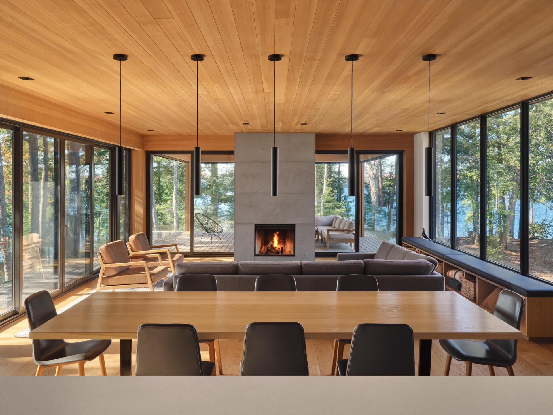 A light-filled living space wrapped in windows and anchored by a sculptural concrete fireplace, with a long built-in bench framing uninterrupted forest views.