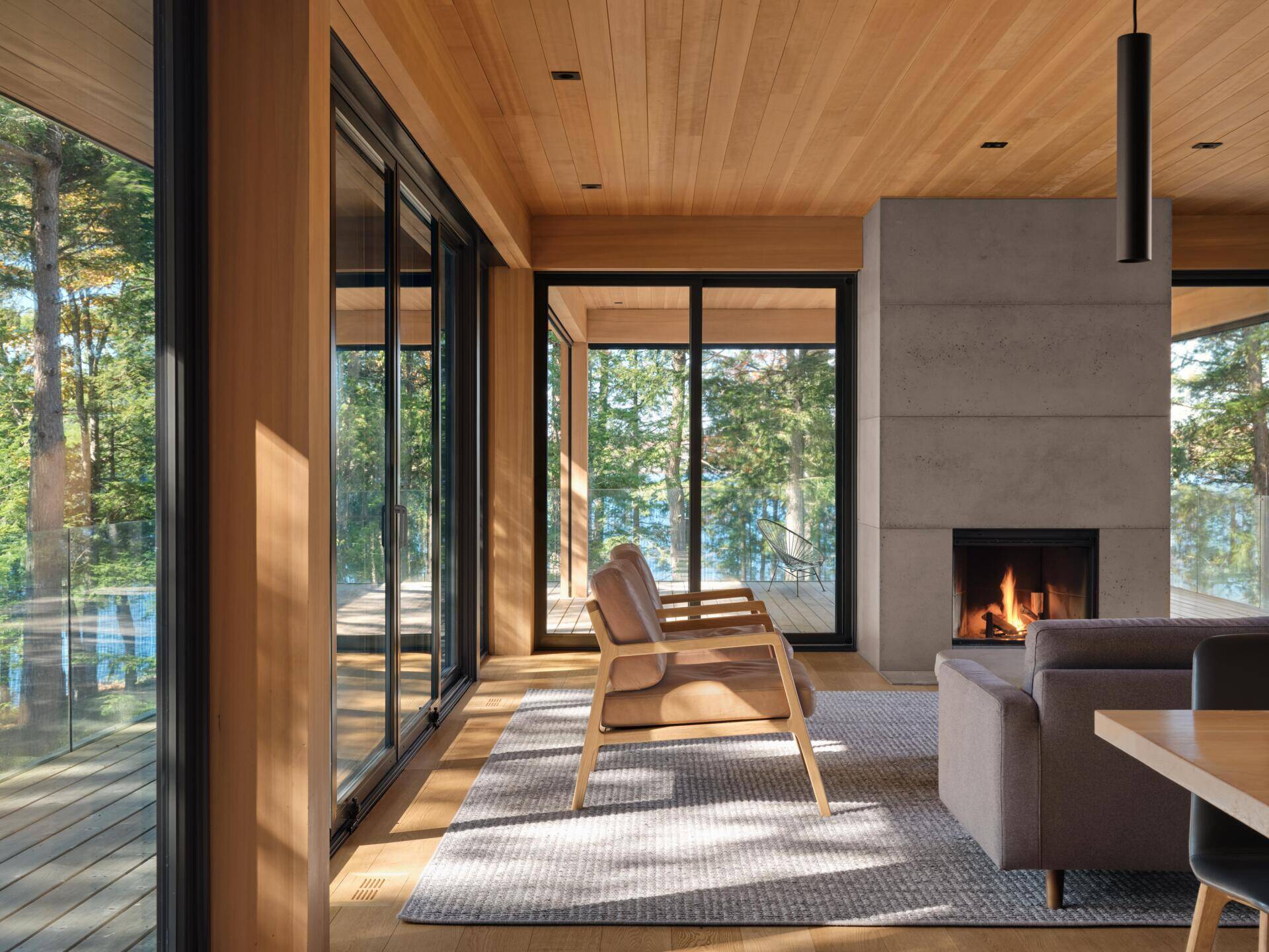 A light-filled living space wrapped in windows and anchored by a sculptural concrete fireplace, with a long built-in bench framing uninterrupted forest views.
