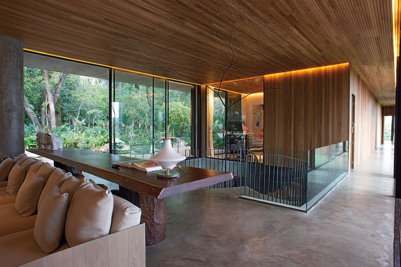 A warm modern interior with a wood ceiling, concrete floors, soft lighting, and a hanging fireplace, balanced by full-height glass walls.