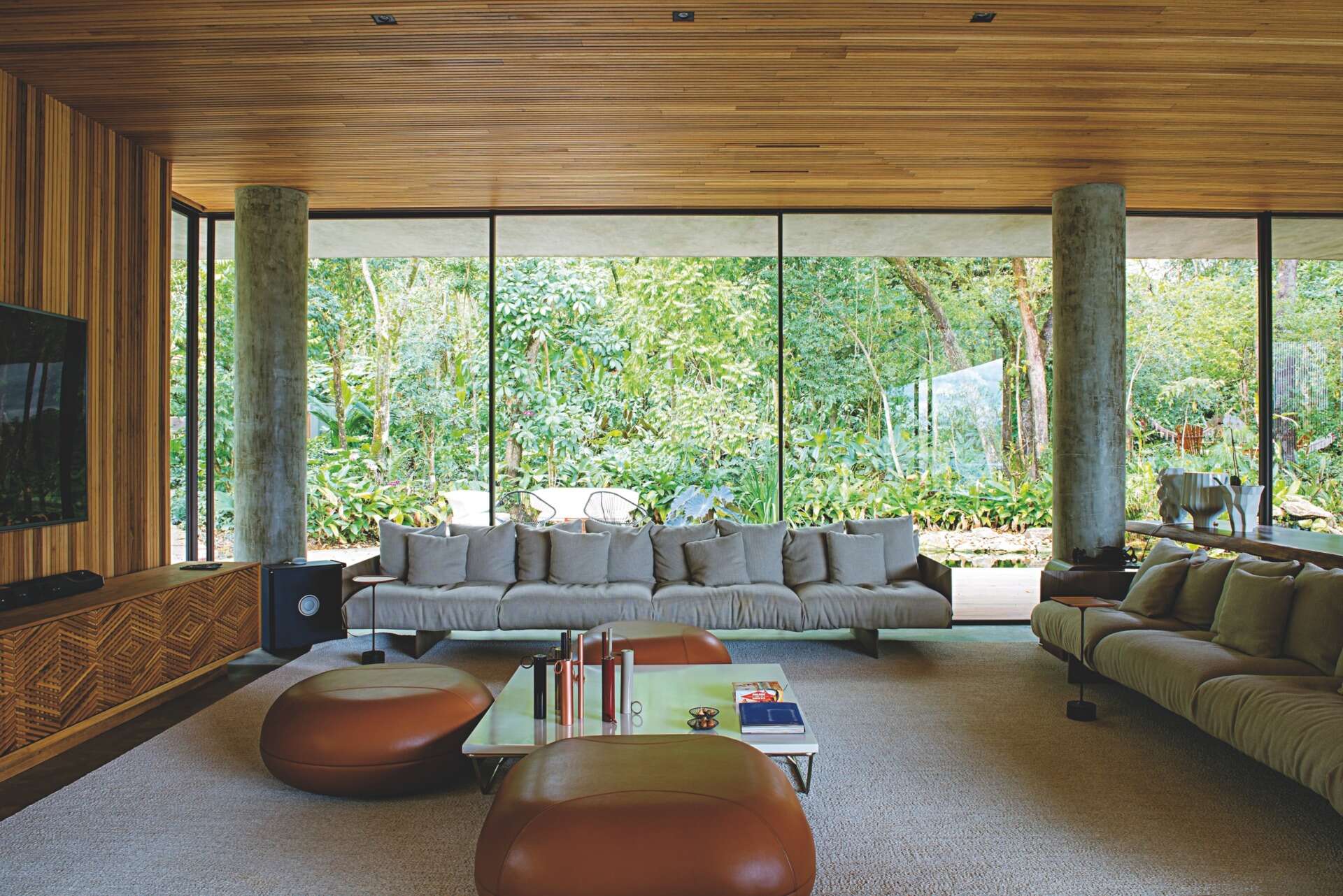 A warm modern interior with a wood ceiling, concrete floors, soft lighting, and a hanging fireplace, balanced by full-height glass walls.