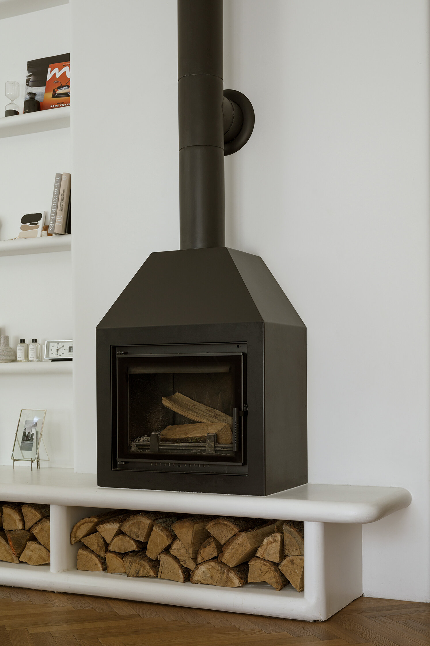 A modern living room with a long, multi-functional unit that integrates the firewood storage while doubling as seating. A built-in shelving niche and fireplace finish the living room space.