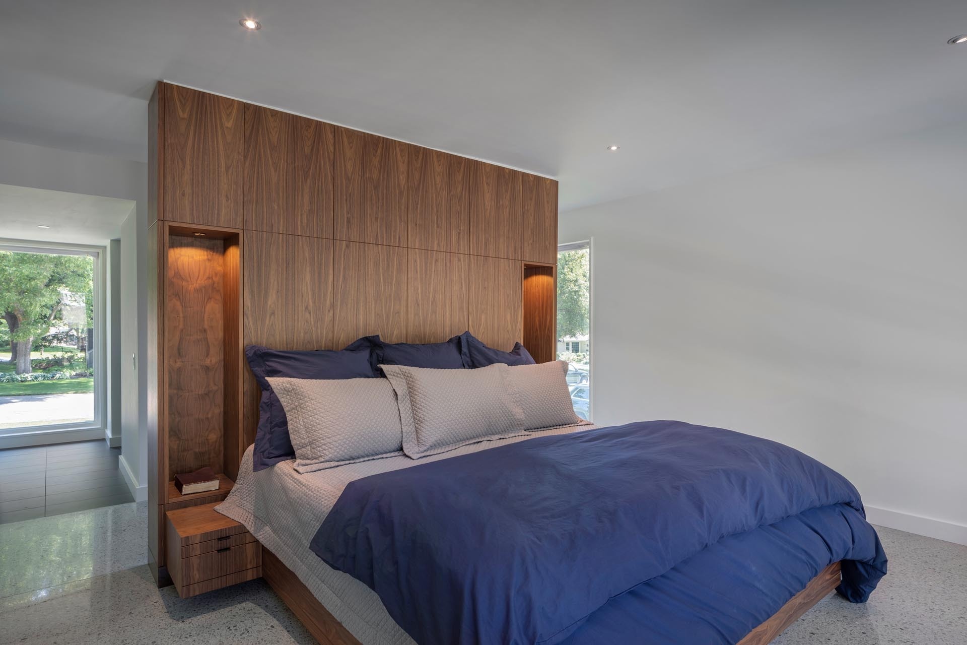 In the primary bedroom, a wood accent wall adds texture and warmth. Carefully placed voids within the wall hold built-in bedside lamps, turning a simple surface into a thoughtful architectural feature.