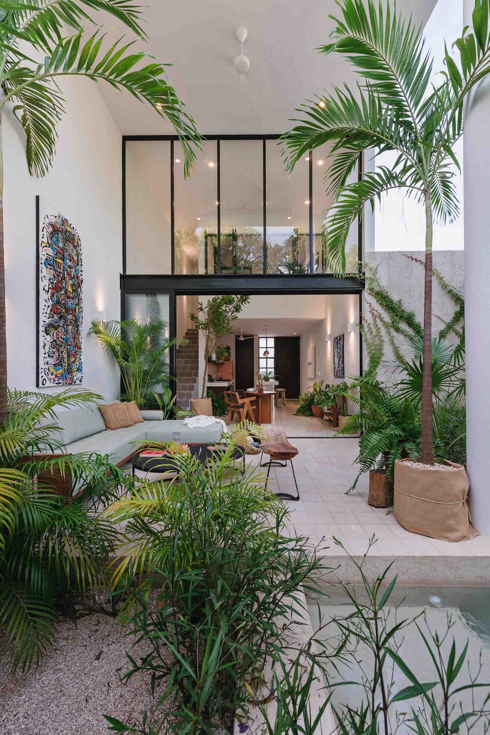 A small courtyard patio filled with tropical plants, colorful artwork, and relaxed seating creates a private outdoor living room inside this modern Mexican home.