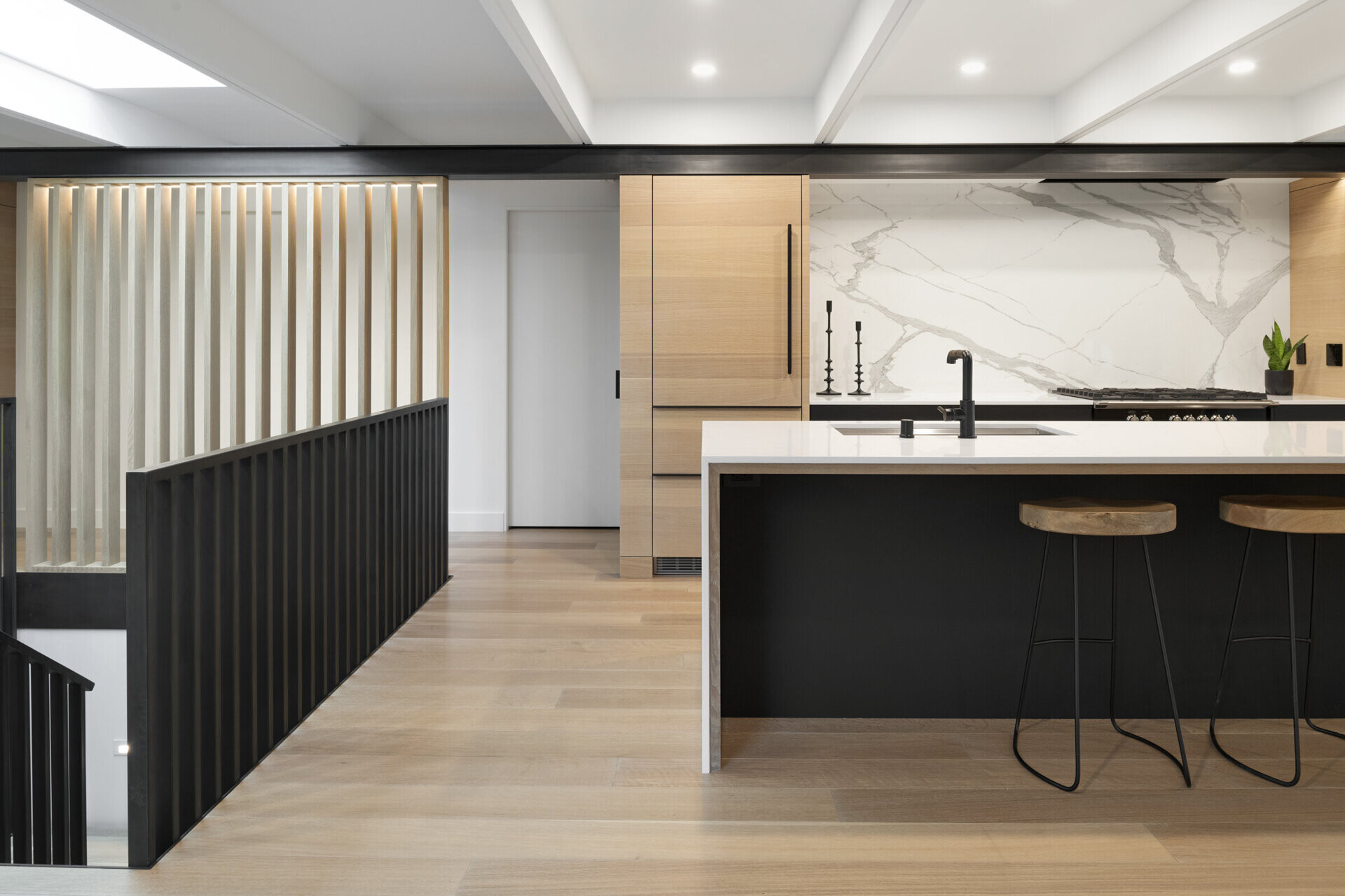 La cocina junto al comedor equilibra gabinetes de madera, herrajes negros simples y encimeras blancas. Ubicada al otro lado de la chimenea de doble cara, crea una sutil conexión visual con el resto de la casa manteniendo su propio espacio único.