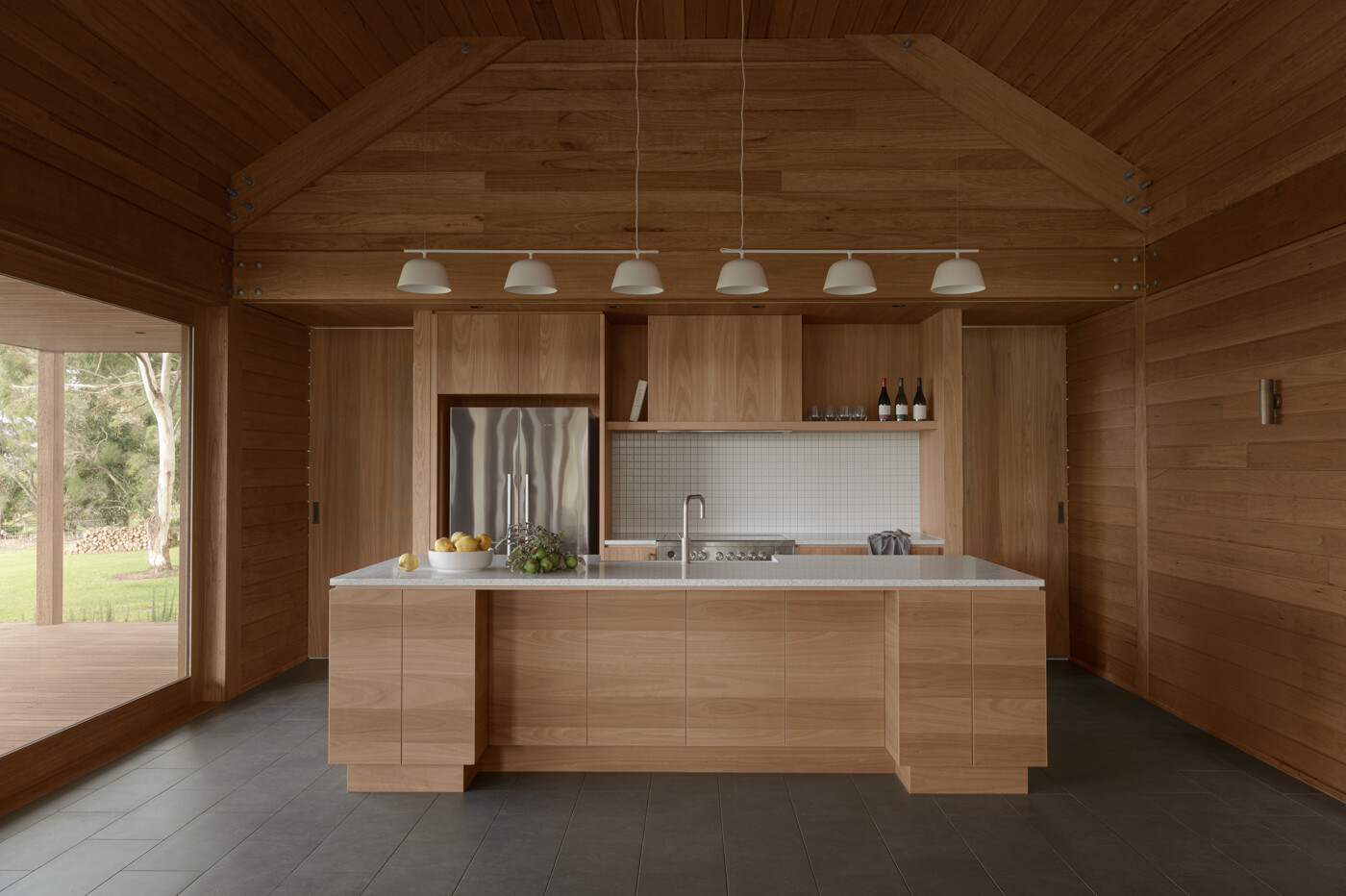 The kitchen sits within the rear pavilion alongside the living area and is positioned to face the vineyard. Wood cabinetry matches the surrounding walls and ceiling, while white counter tops, lighting, and backsplash provide a contrasting element.