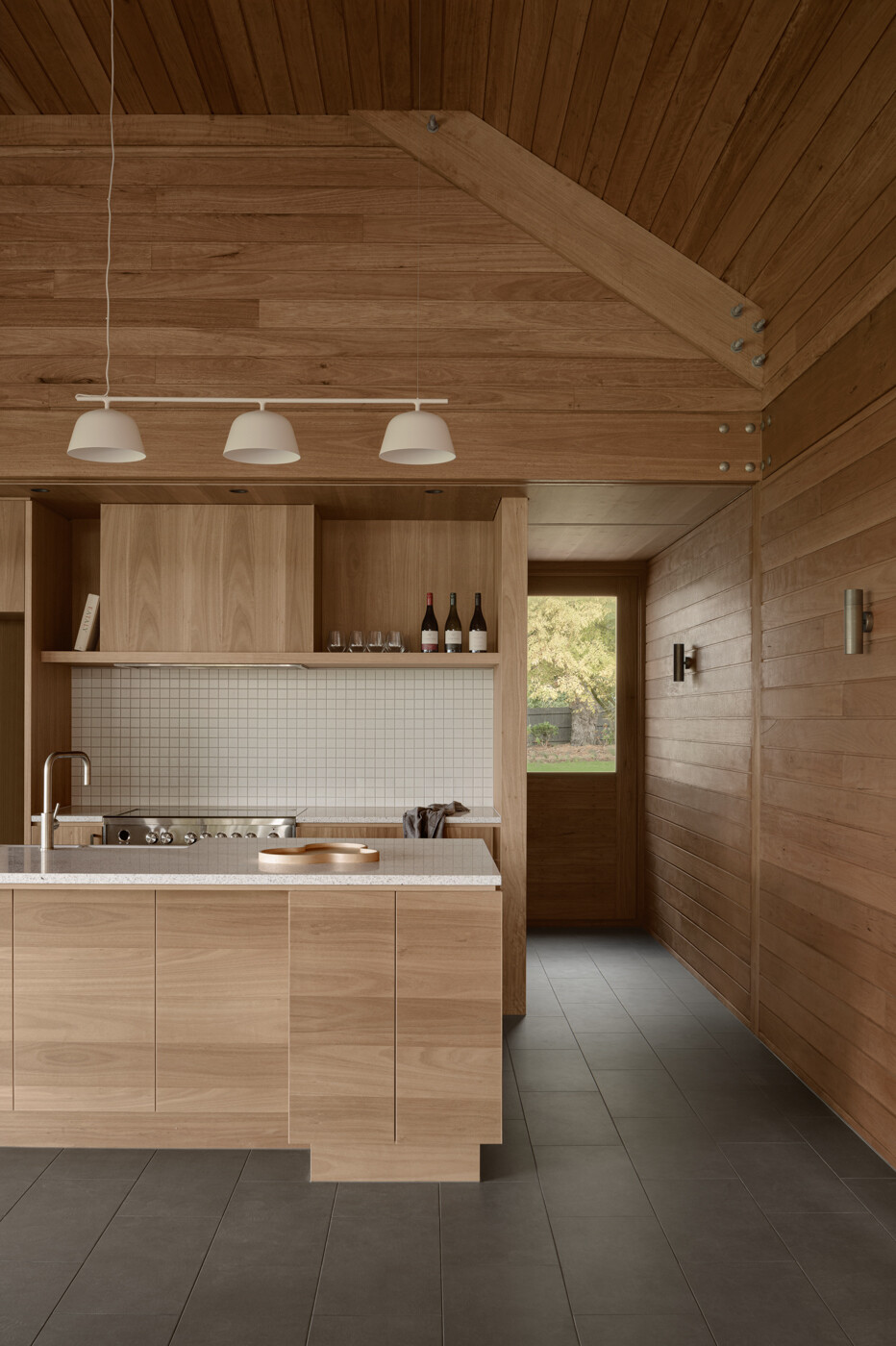 The kitchen sits within the rear pavilion alongside the living area and is positioned to face the vineyard. Wood cabinetry matches the surrounding walls and ceiling, while white counter tops, lighting, and backsplash provide a contrasting element.