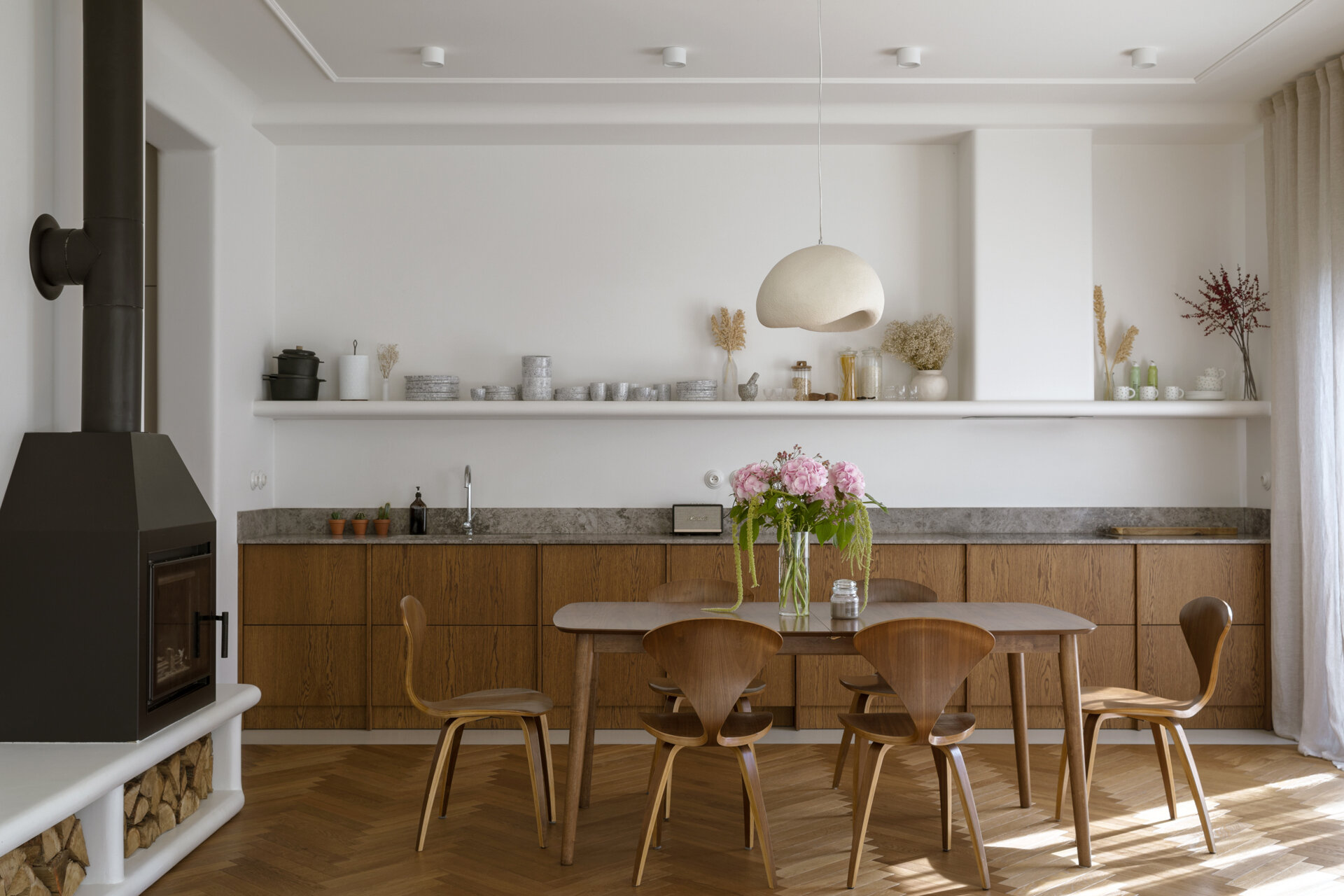 The dining room separates the kitchen from the living room. A concealed storage area keeps the space visually clean, allowing materials like the wood cabinets, and form to take focus. A shelf with a curved front creates a place to display kitchenware.