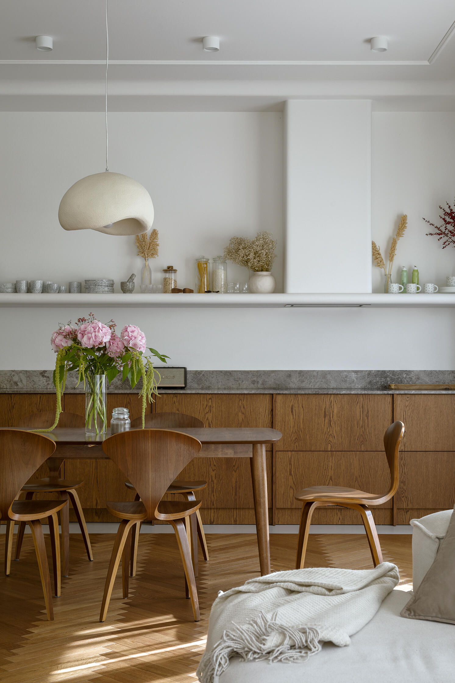 The dining room separates the kitchen from the living room. A concealed storage area keeps the space visually clean, allowing materials like the wood cabinets, and form to take focus. A shelf with a curved front creates a place to display kitchenware.