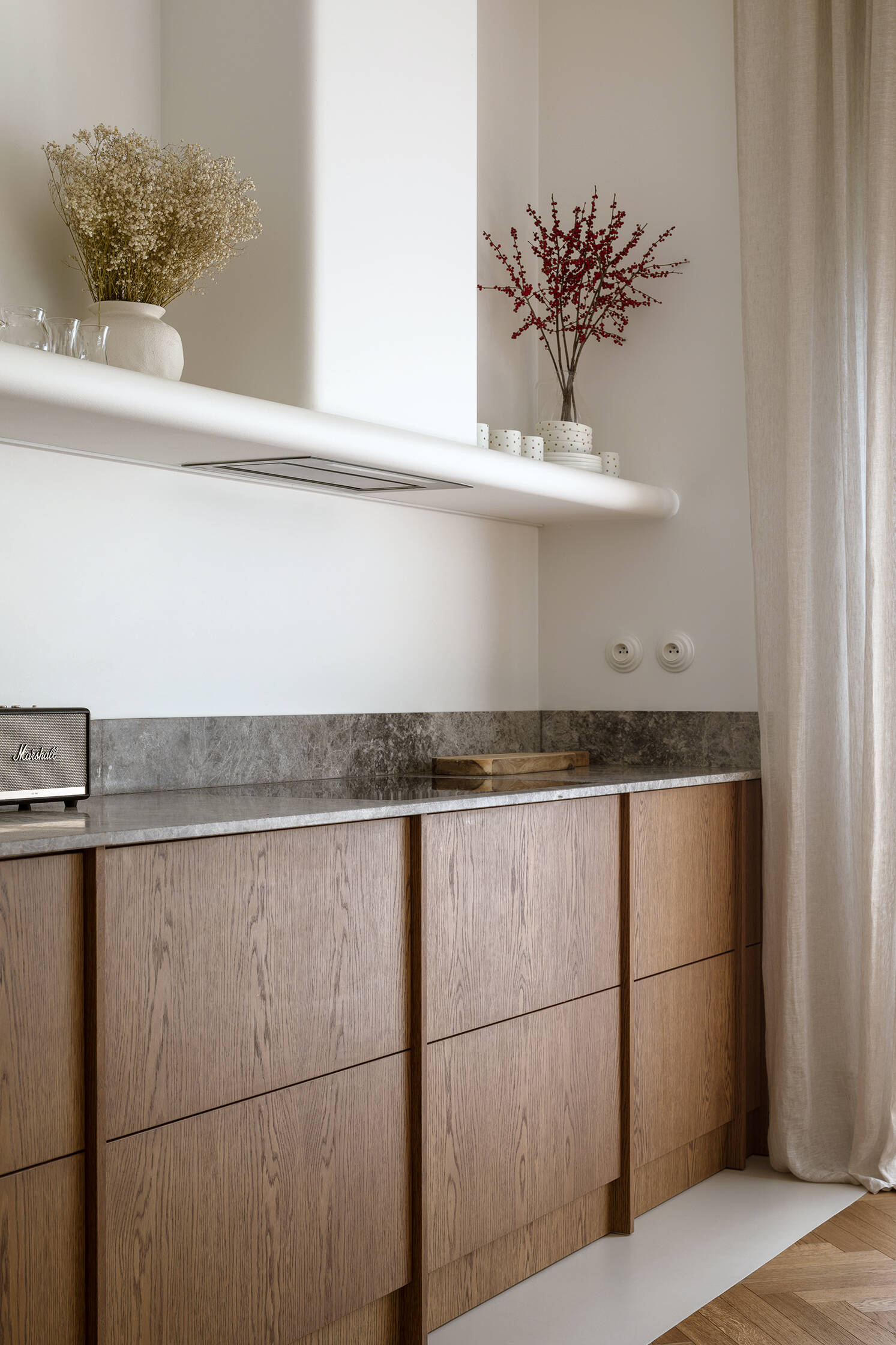 The dining room separates the kitchen from the living room. A concealed storage area keeps the space visually clean, allowing materials like the wood cabinets, and form to take focus. A shelf with a curved front creates a place to display kitchenware.
