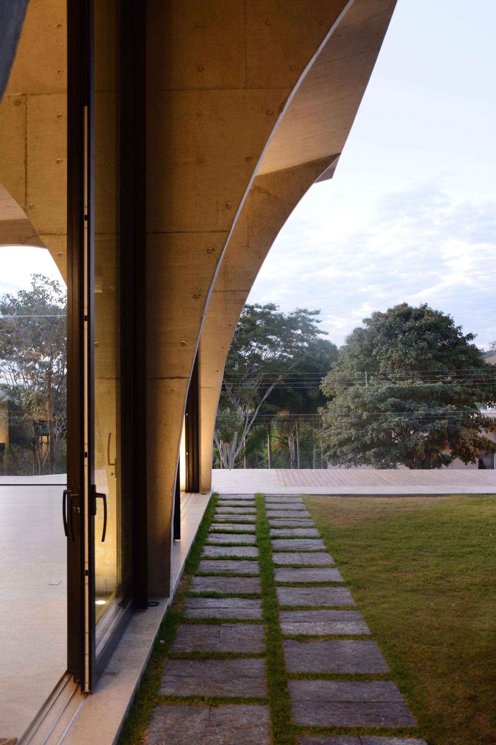 The living room, dining area, and kitchen share a dramatic double height space framed by concrete arches and large sliding glass doors that connect the interior to the gardens.
