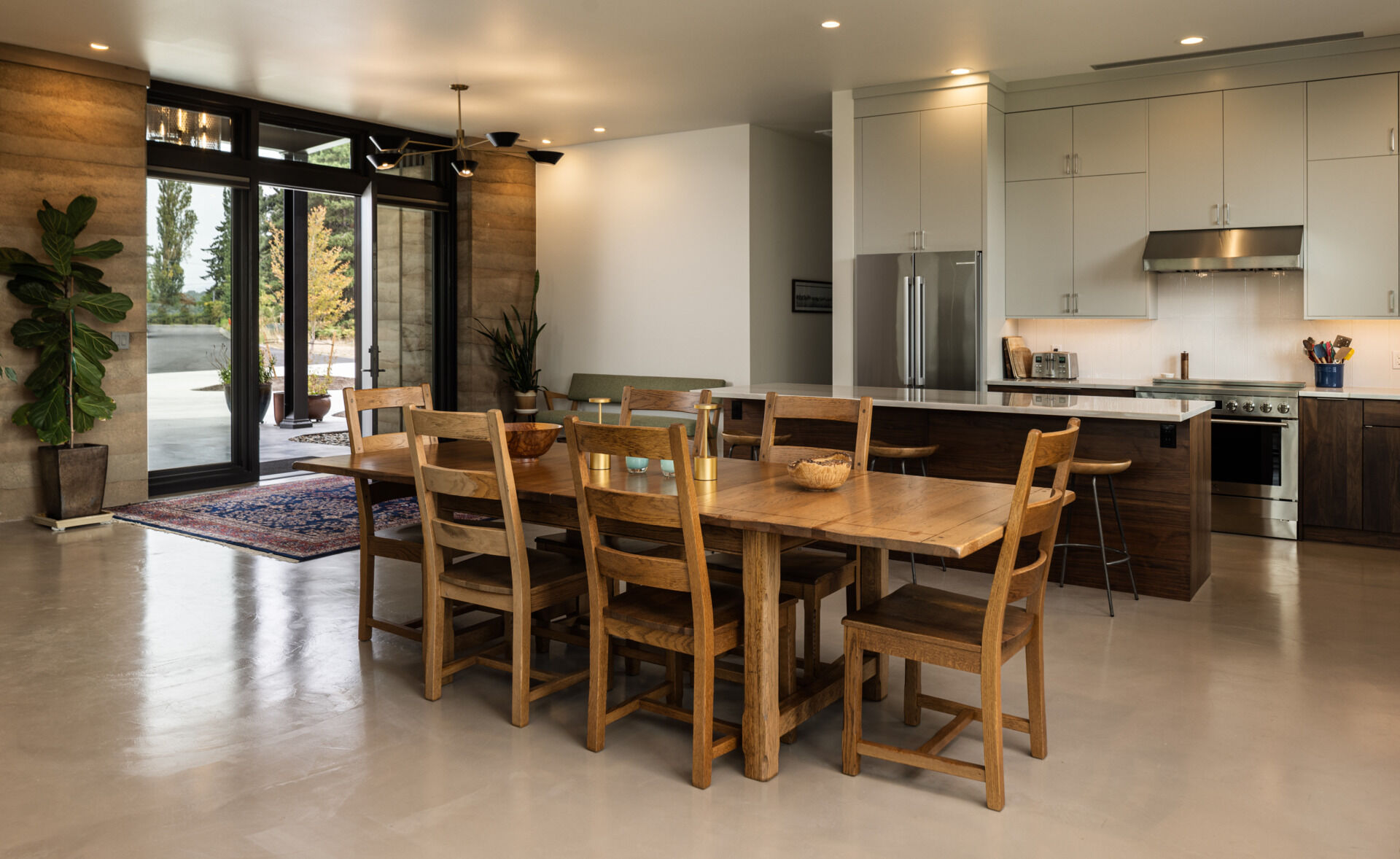 The great room combines the kitchen, dining area, and sitting space in an open layout framed by sliding glass doors and textured rammed earth walls.