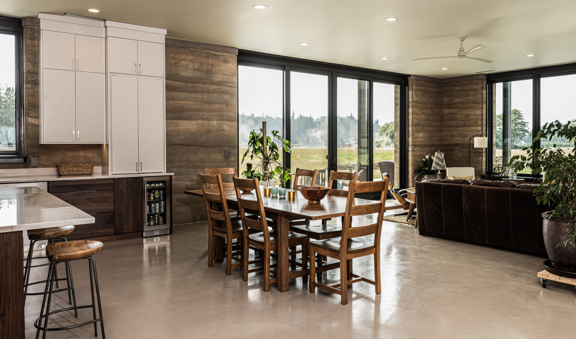 The great room combines the kitchen, dining area, and sitting space in an open layout framed by sliding glass doors and textured rammed earth walls.