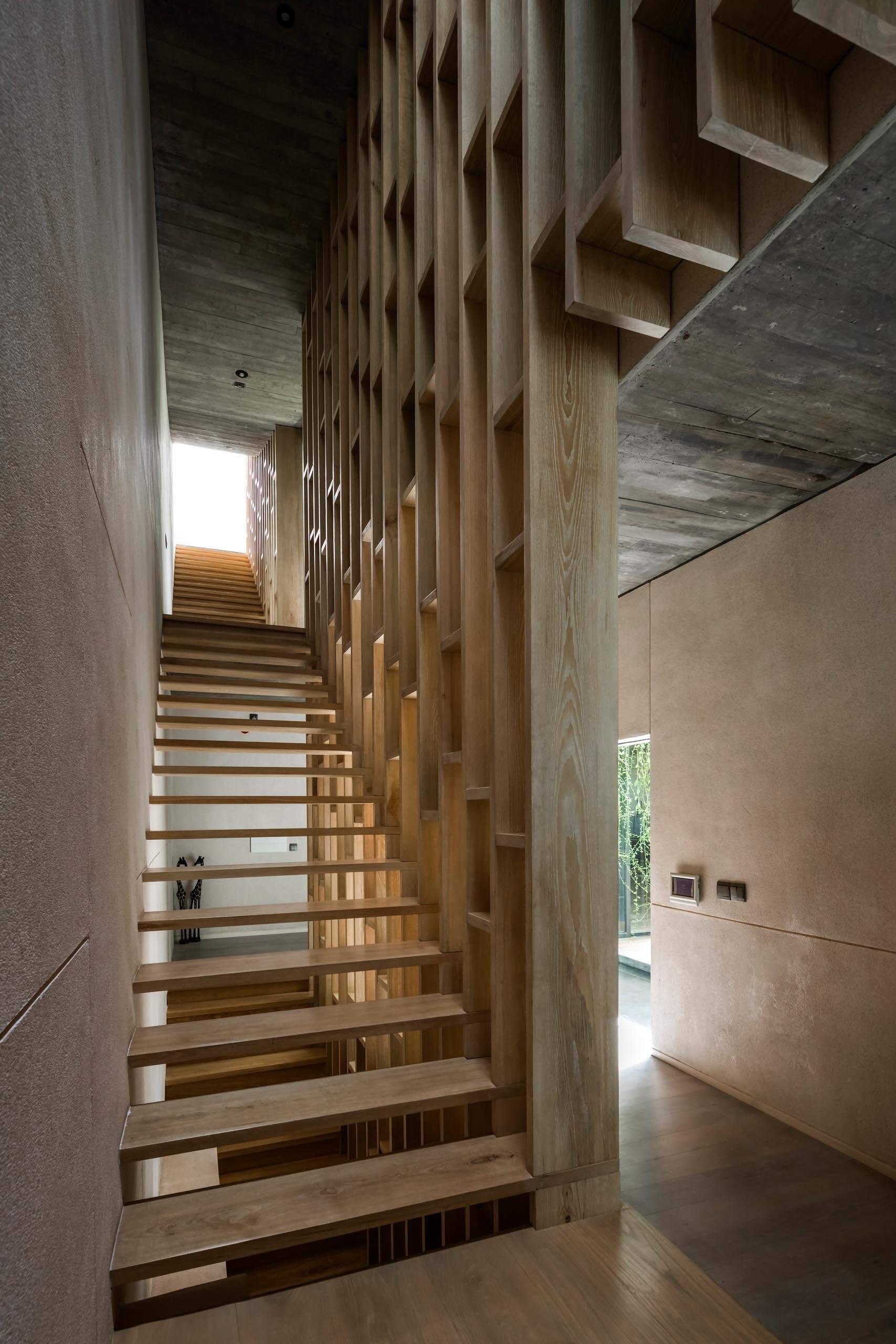 Las paredes de hormigón en bruto, las cálidas escaleras de madera y la luz filtrada crean un ambiente interior relajante, con plantas y materiales naturales que suavizan la arquitectura.