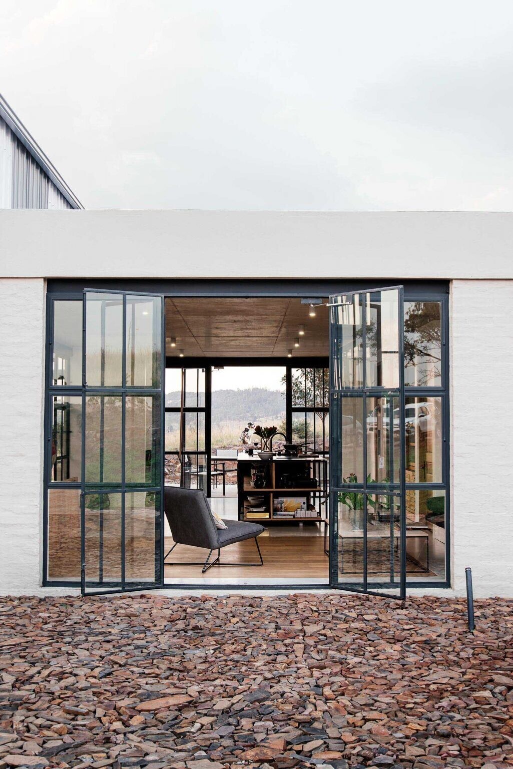 Black-framed glass doors connect the conservatory to the kitchen, where a black island becomes the focal point. Open wood shelving beneath it displays everyday items like pots, pans, and serving dishes, turning storage into part of the design.
