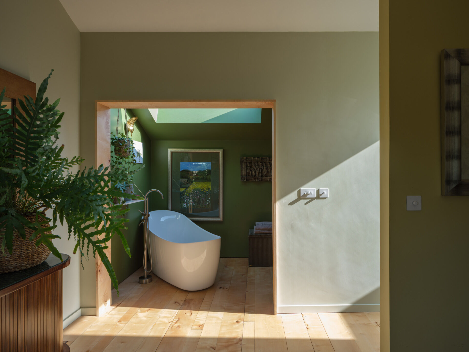 A freestanding tub set within a green alcove, skylit from above and softened with timber floors and plants.