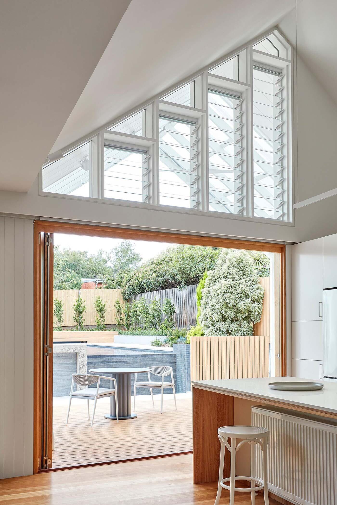 A clean, modern kitchen pairs white cabinetry with timber accents, opening directly to outdoor dining and supported by hidden functional spaces.