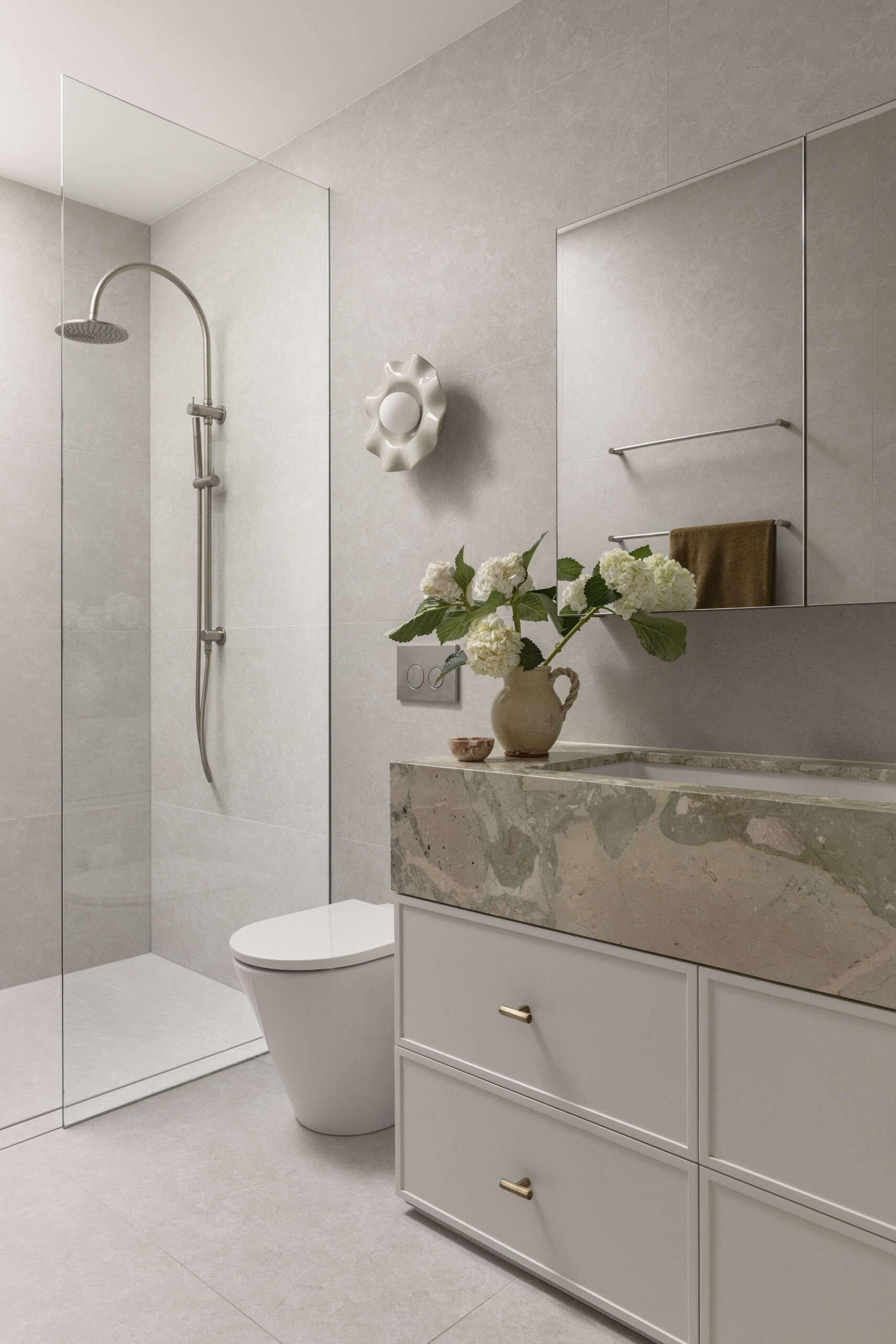 This primary en-suite bathroom has soft stone-look surfaces, a frameless glass shower, and warm brass details bringing subtle polish to the room. The veined vanity surround adds just enough movement and color, while the sculptural wall light and fresh white blooms keep the space feeling personal and calm.