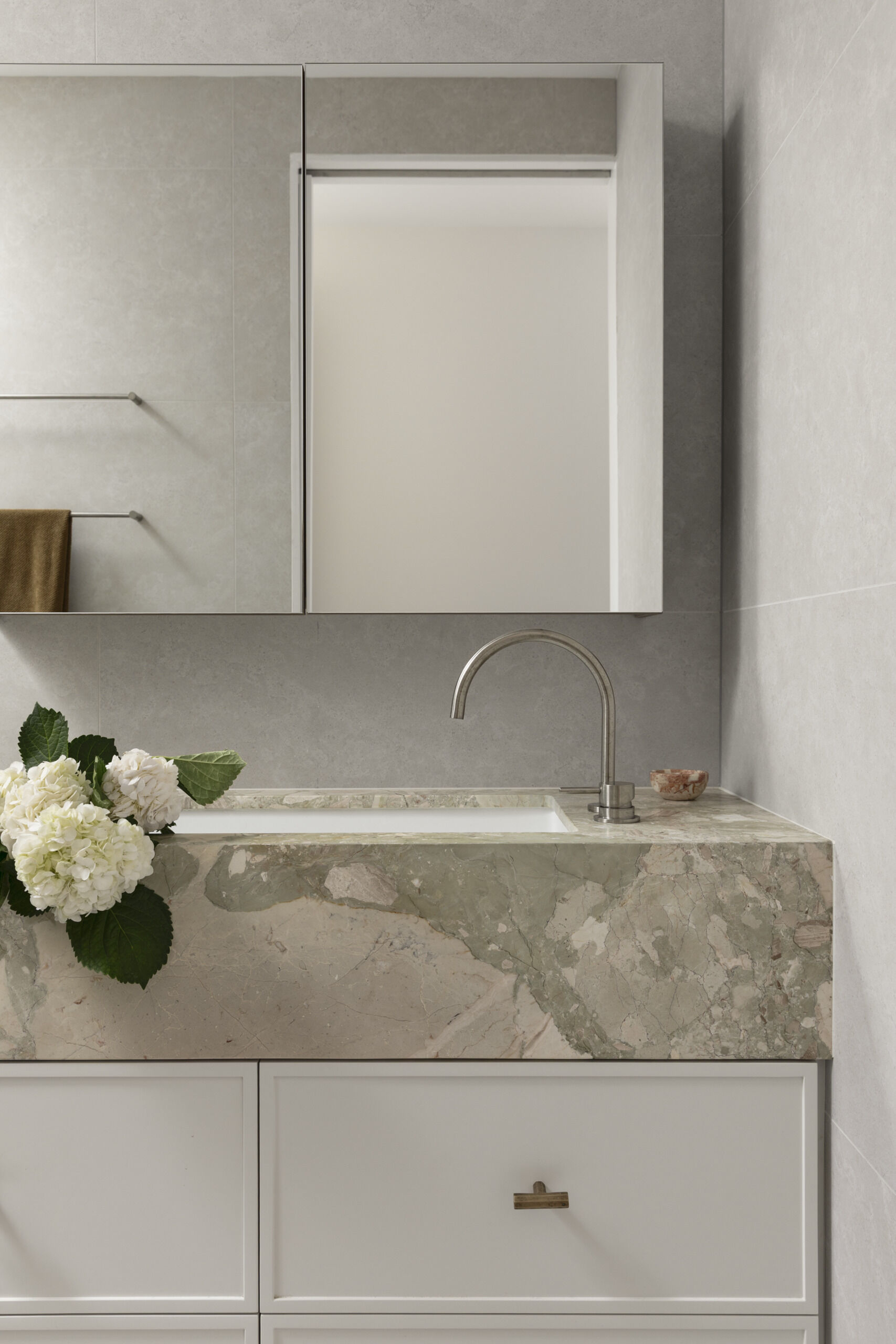 This primary en-suite bathroom has soft stone-look surfaces, a frameless glass shower, and warm brass details bringing subtle polish to the room. The veined vanity surround adds just enough movement and color, while the sculptural wall light and fresh white blooms keep the space feeling personal and calm.