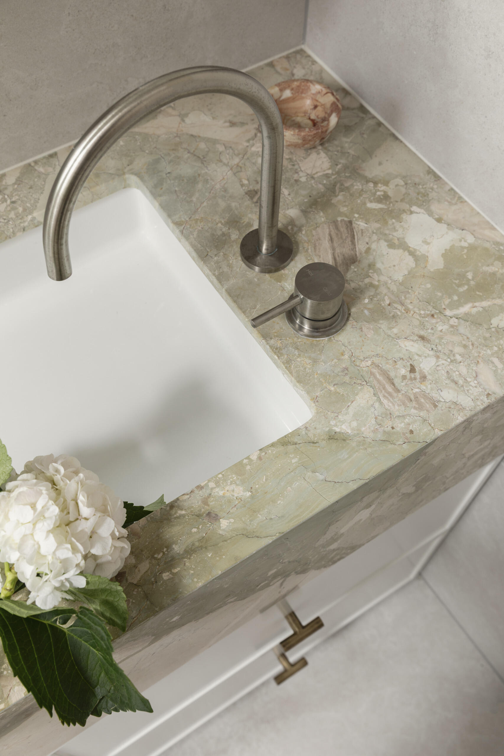 This primary en-suite bathroom has soft stone-look surfaces, a frameless glass shower, and warm brass details bringing subtle polish to the room. The veined vanity surround adds just enough movement and color, while the sculptural wall light and fresh white blooms keep the space feeling personal and calm.