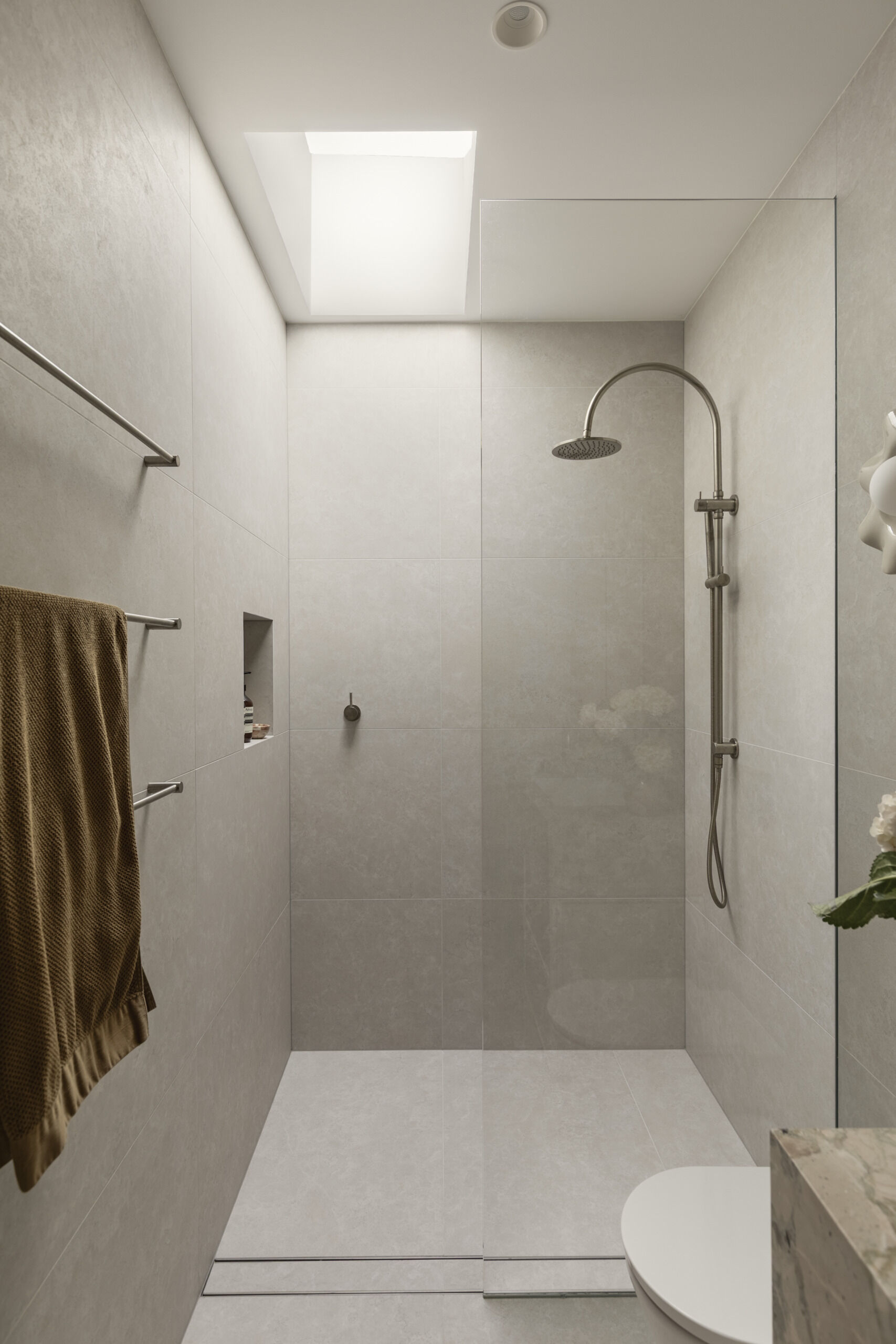This primary en-suite bathroom has soft stone-look surfaces, a frameless glass shower, and warm brass details bringing subtle polish to the room. The veined vanity surround adds just enough movement and color, while the sculptural wall light and fresh white blooms keep the space feeling personal and calm.