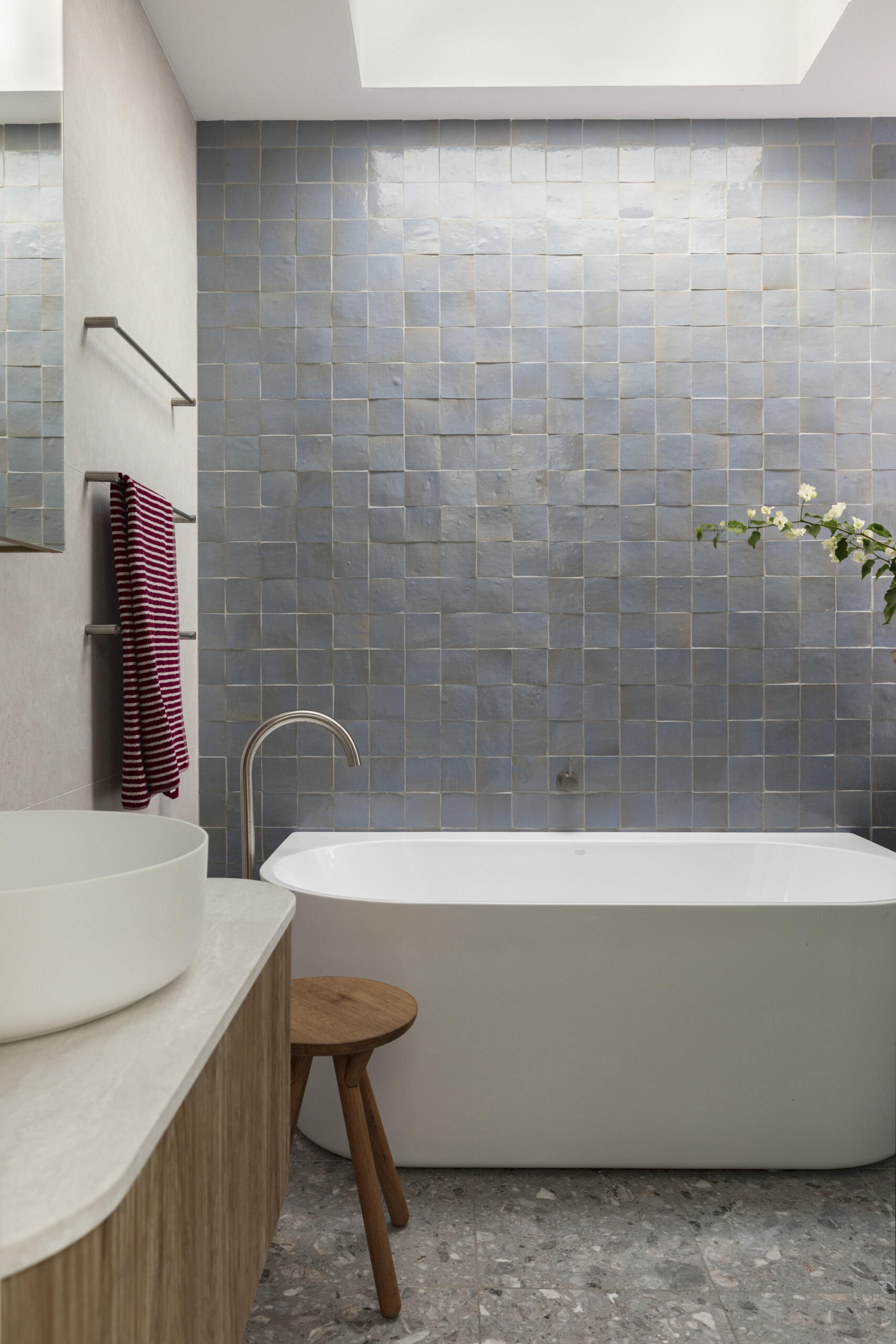The upstairs family bathroom features soft gray walls, cloudy blue tiles, and a crisp white tub creating a fresh, airy look under the skylight. The light wood vanity and small stool bring in warmth, while the terrazzo floor adds texture and everyday durability.