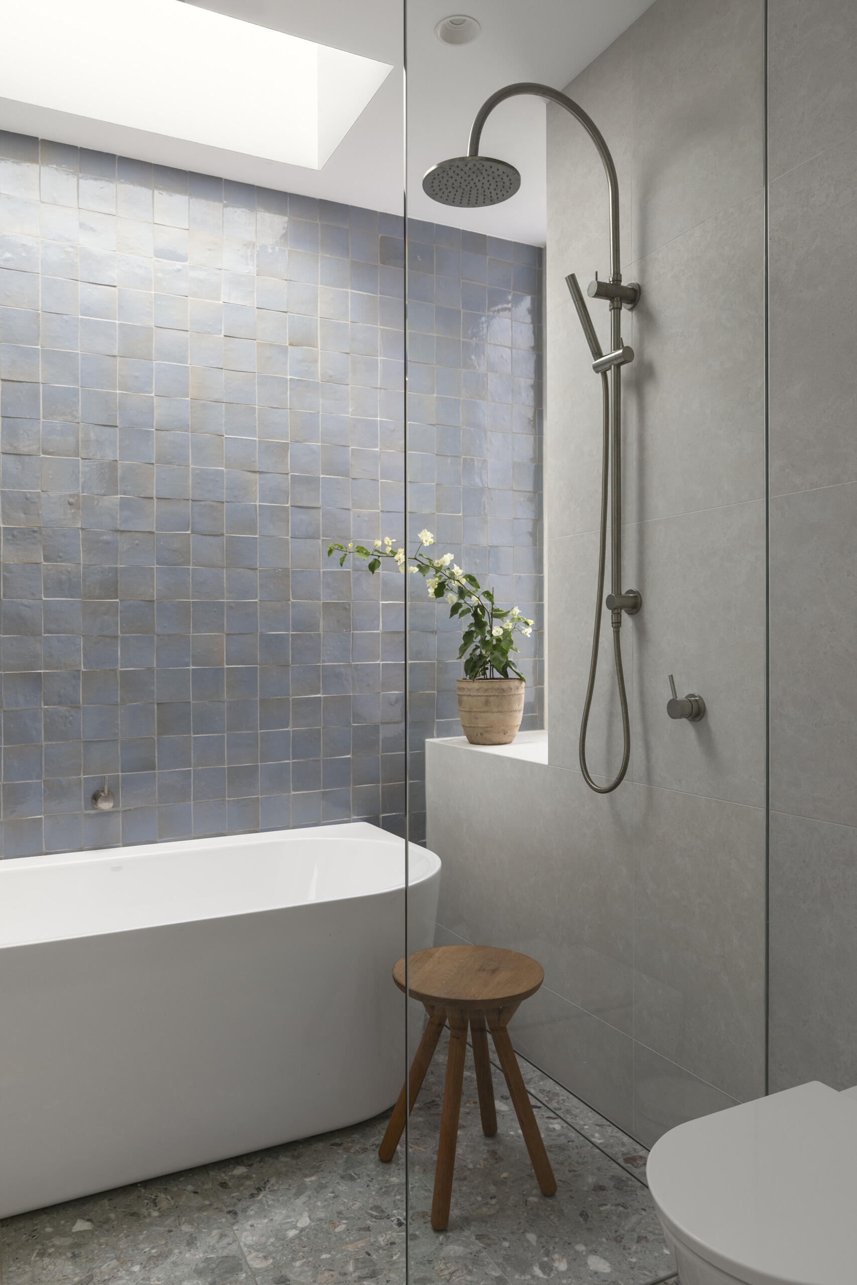 The upstairs family bathroom features soft gray walls, cloudy blue tiles, and a crisp white tub creating a fresh, airy look under the skylight. The light wood vanity and small stool bring in warmth, while the terrazzo floor adds texture and everyday durability.