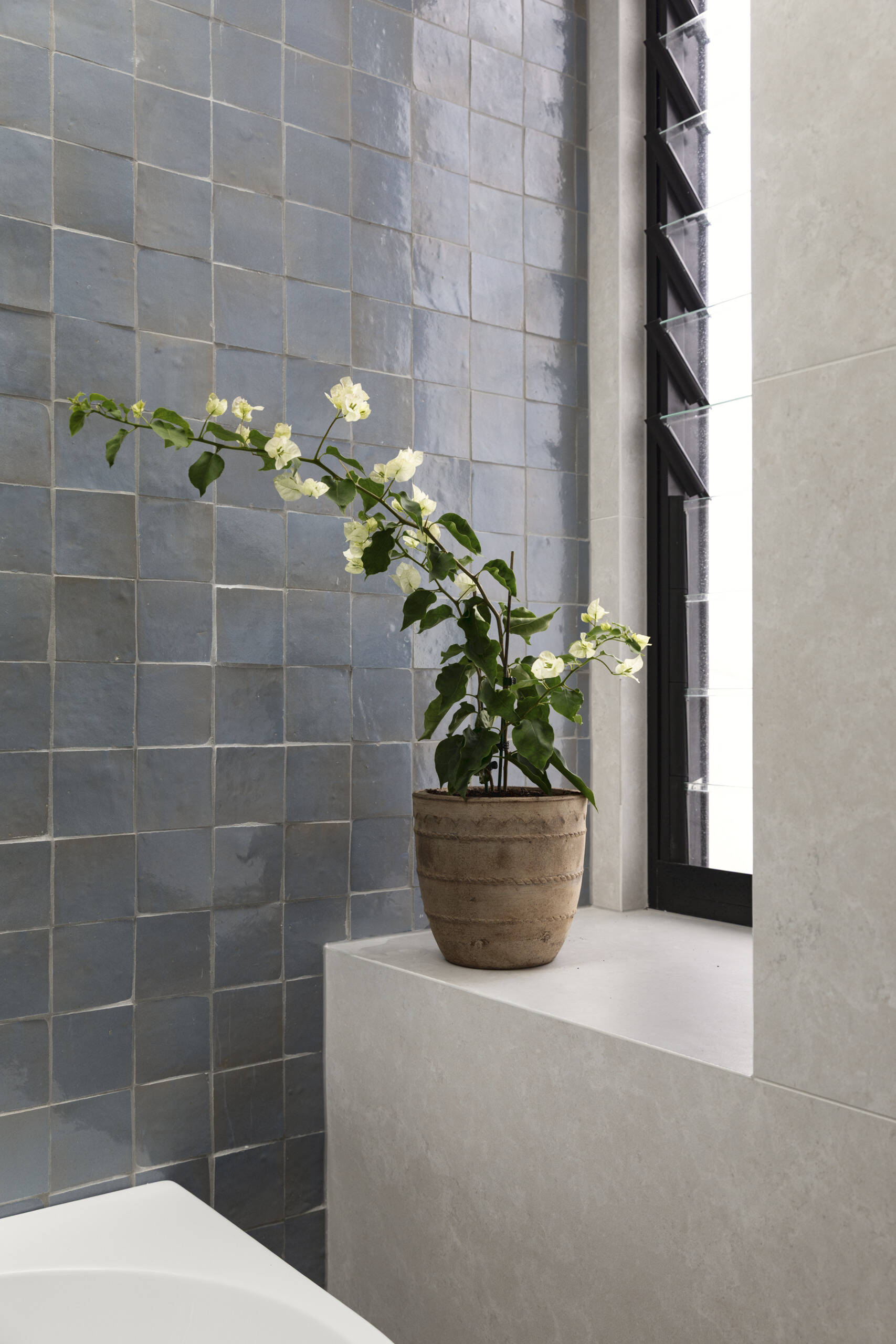 The upstairs family bathroom features soft gray walls, cloudy blue tiles, and a crisp white tub creating a fresh, airy look under the skylight. The light wood vanity and small stool bring in warmth, while the terrazzo floor adds texture and everyday durability.