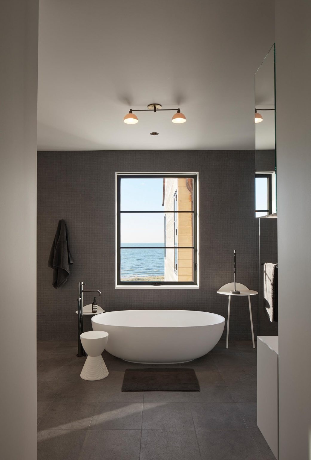 The primary bathroom continues the restrained palette. A freestanding bathtub sits beneath a window, while dark gray walls pair with gray floor tiles to create a cohesive, calming environment.