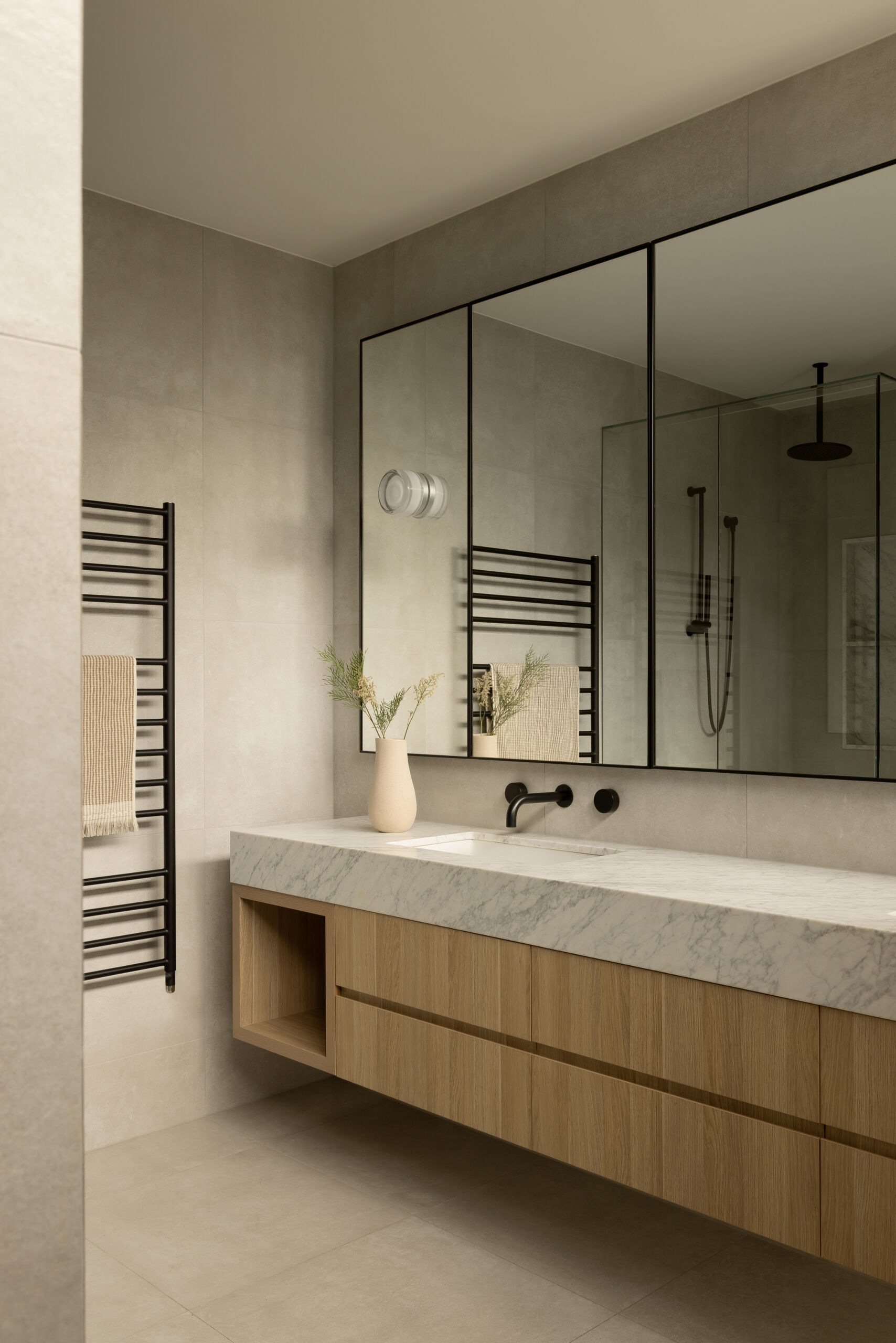 A warm, modern bathroom with a floating timber vanity, marble countertop, and soft neutral tiles, finished with black fixtures for contrast.