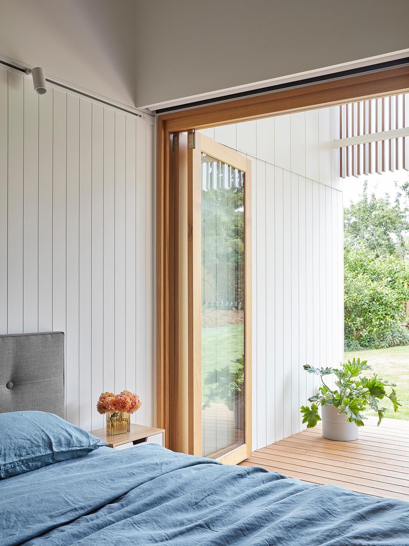 A tucked-away bedroom combines grooved white walls, folding glass doors, and direct deck access for a calm and flexible private space.