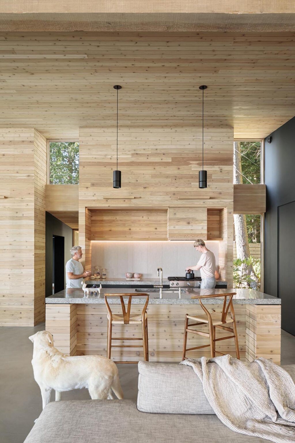 A dining space with a wood feature wall and concrete floors flows into a kitchen with thick countertops and dark cabinetry.