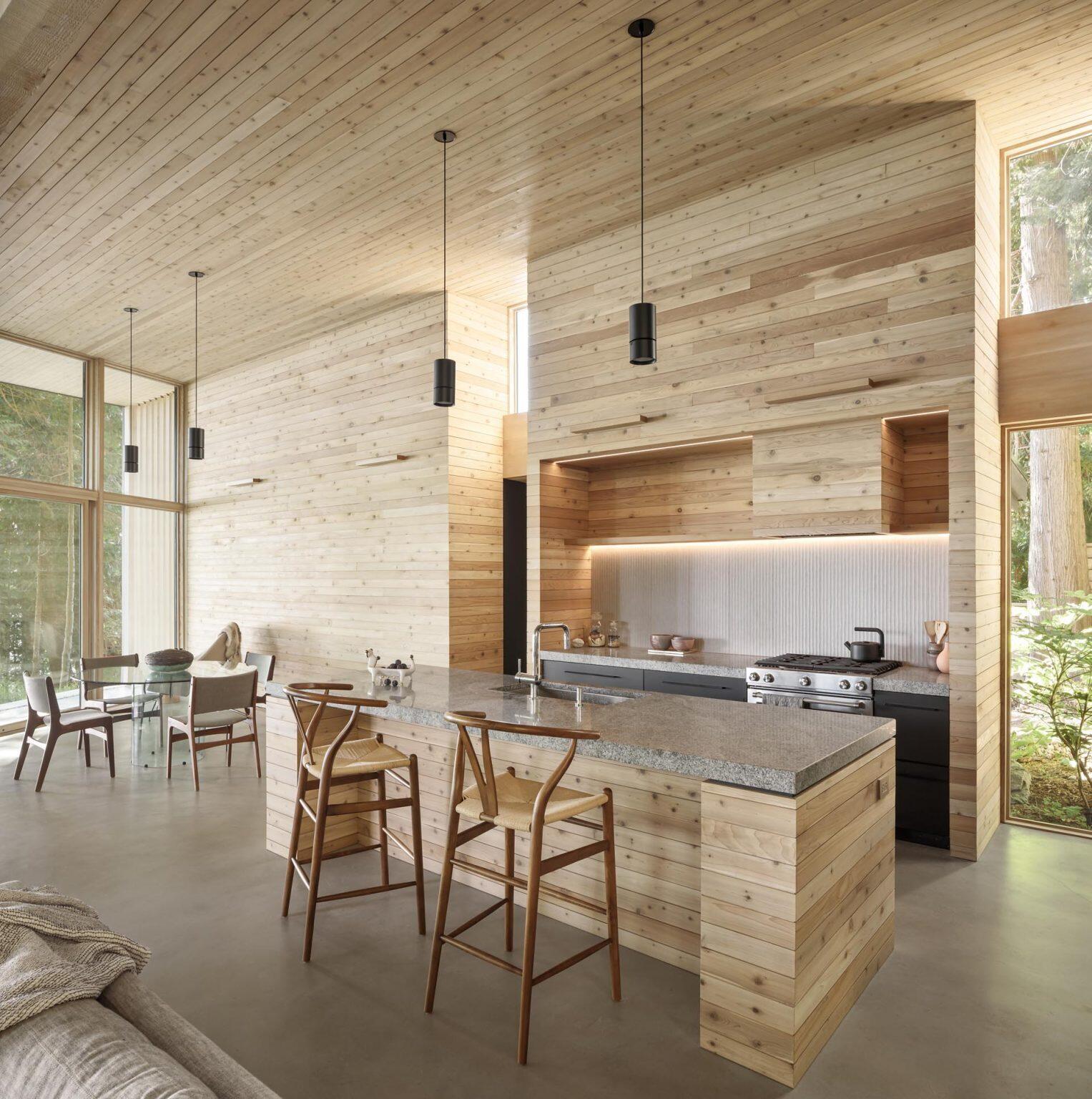 A dining space with a wood feature wall and concrete floors flows into a kitchen with thick countertops and dark cabinetry.