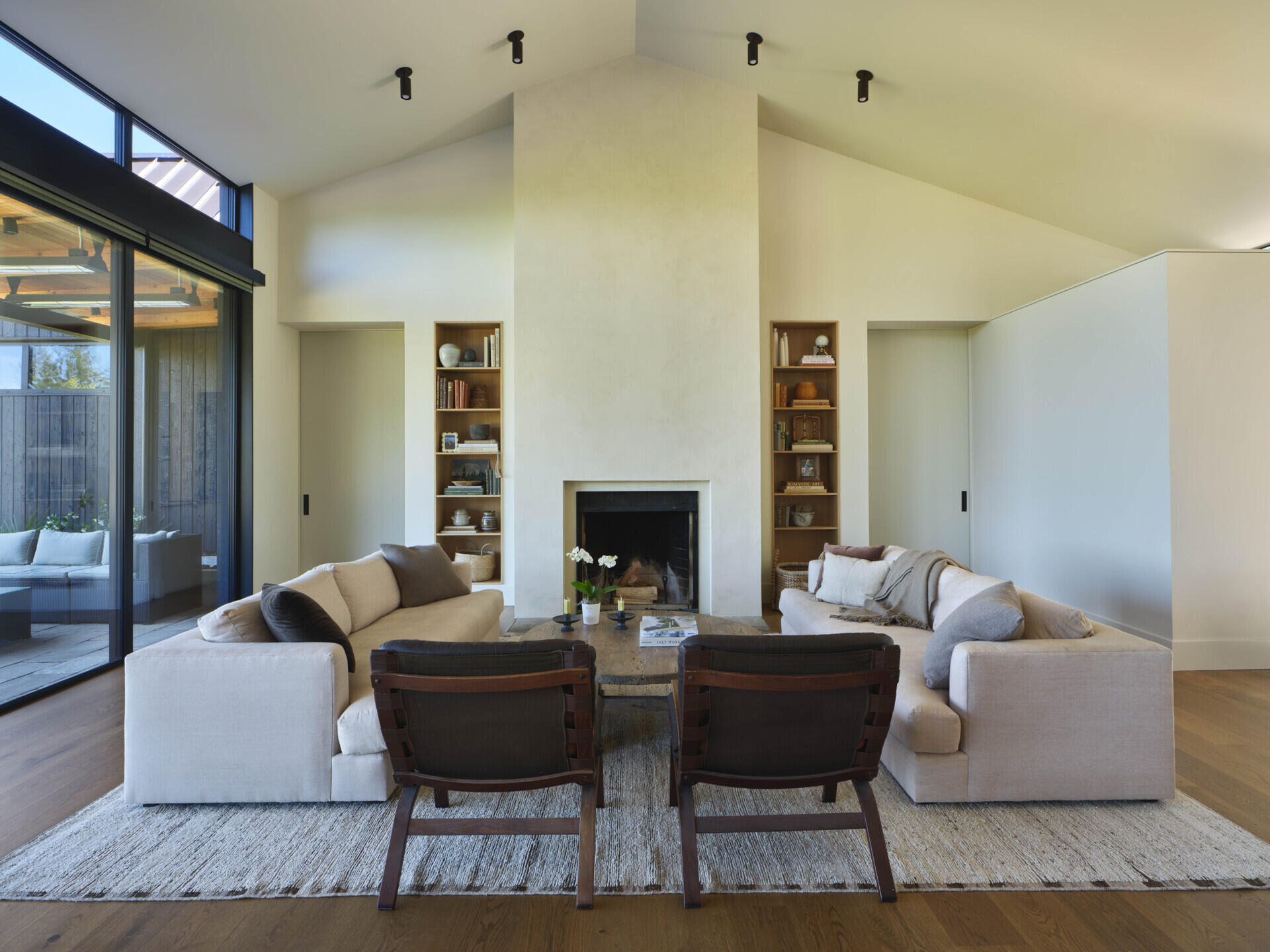 An open living space with large sliding doors connects directly to a covered patio for easy indoor-outdoor flow. A wood-burning fireplace ties into the central chimney volume, adding warmth both visually and physically.