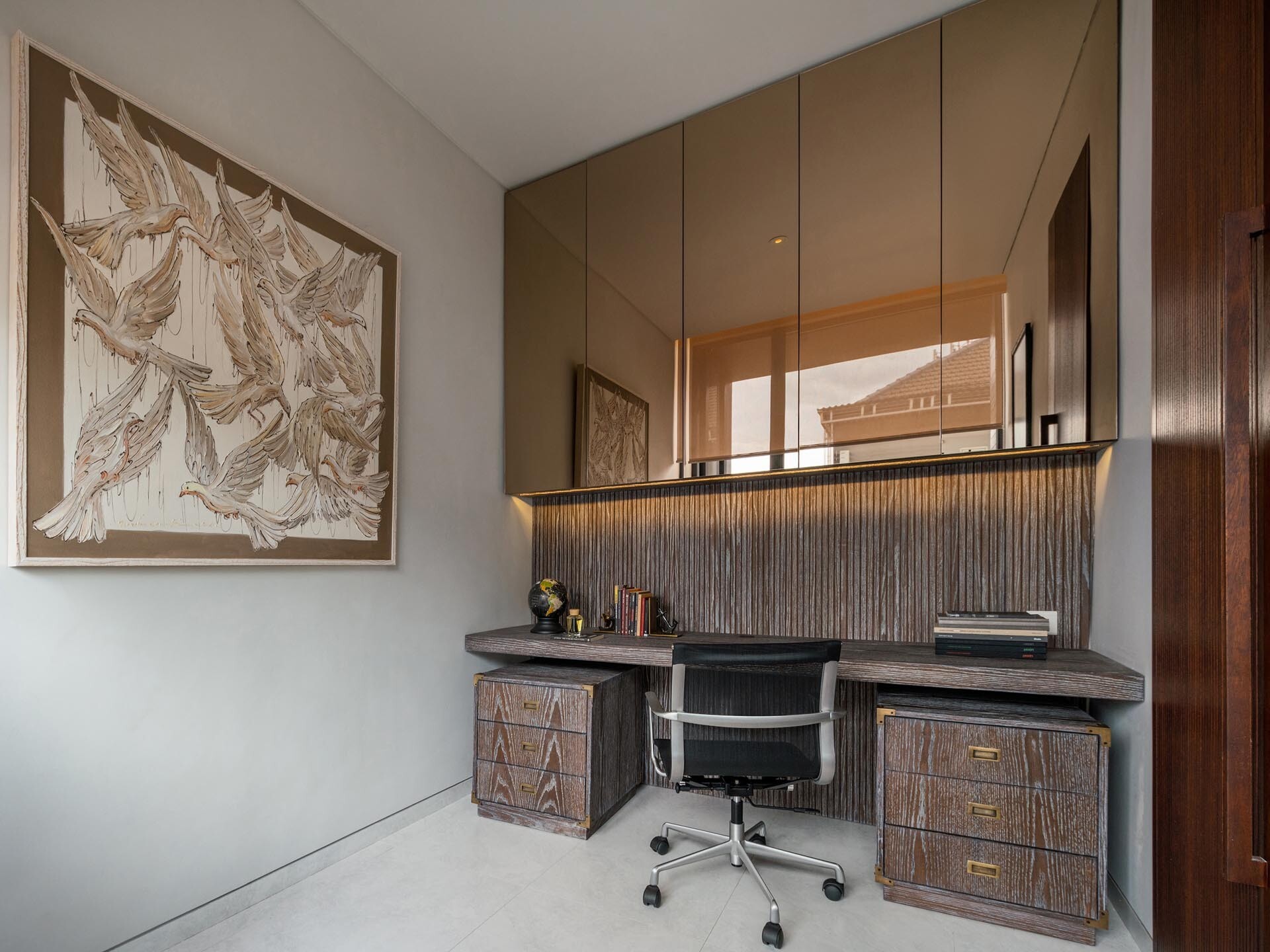 A home office is fitted with mirrored cabinets above the desk, helping reflect light and visually expand the space. A chair placed by the window creates a comfortable corner for breaks or reading.