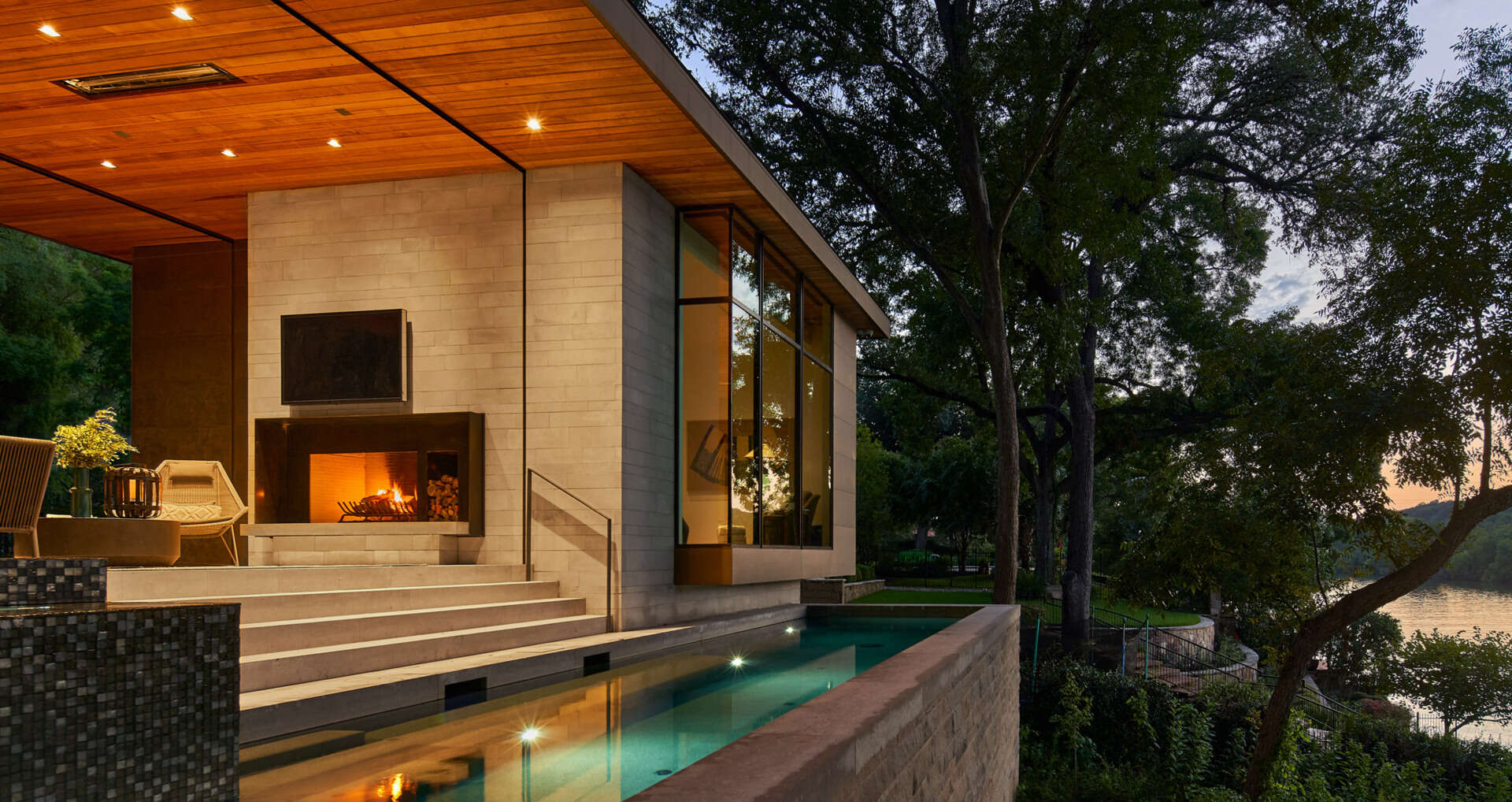 A covered outdoor area with lounge seating, dining space, and a fireplace connects the home to a pool overlooking the lake.
