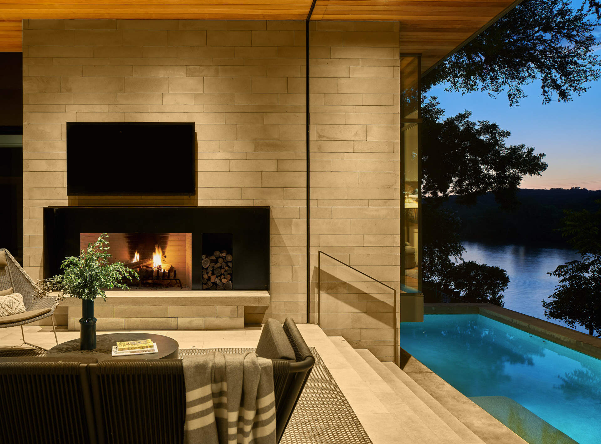 A covered outdoor area with lounge seating, dining space, and a fireplace connects the home to a pool overlooking the lake.