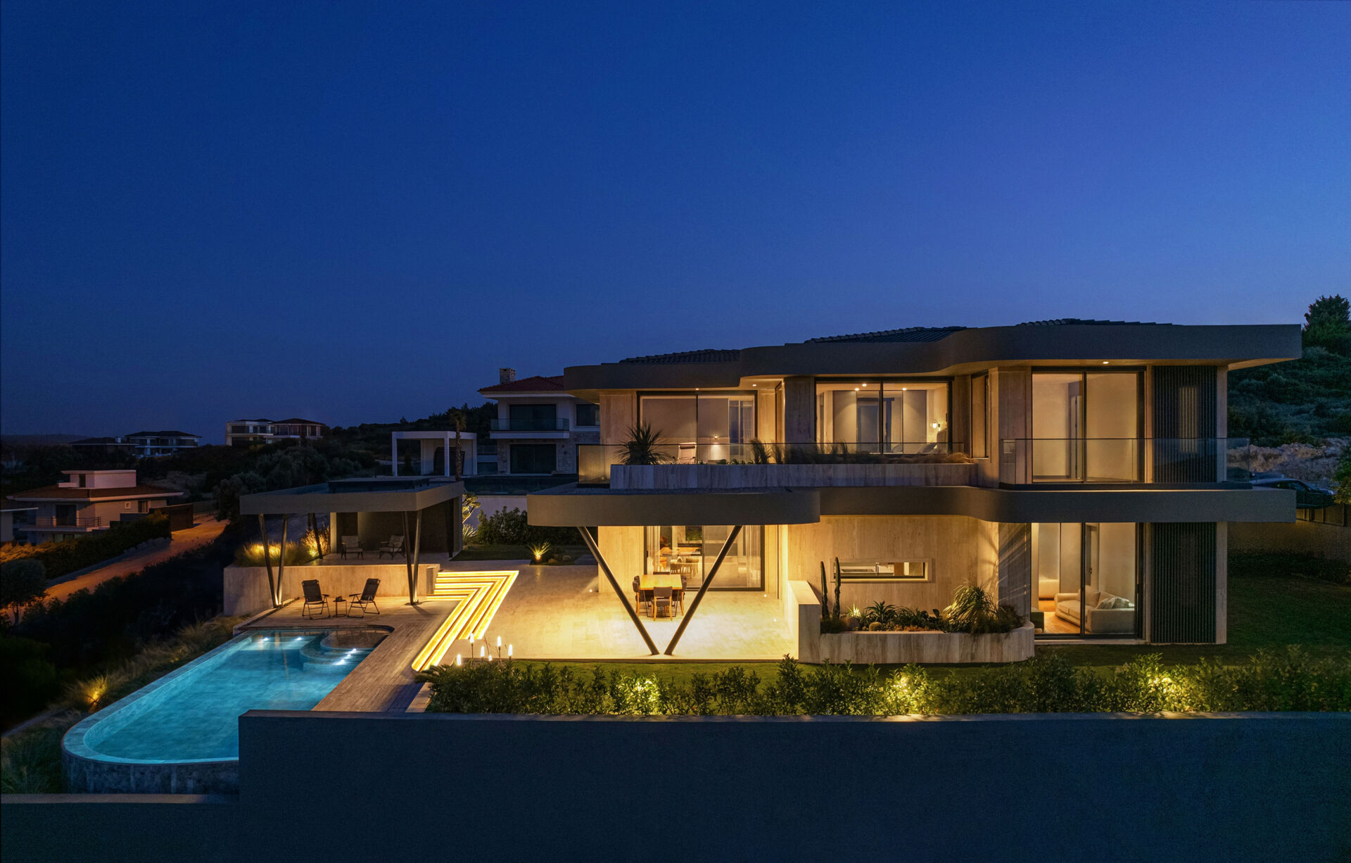 A modern coastal home glowing at night, with soft lighting highlighting sculptural eaves and reflecting across the pool.