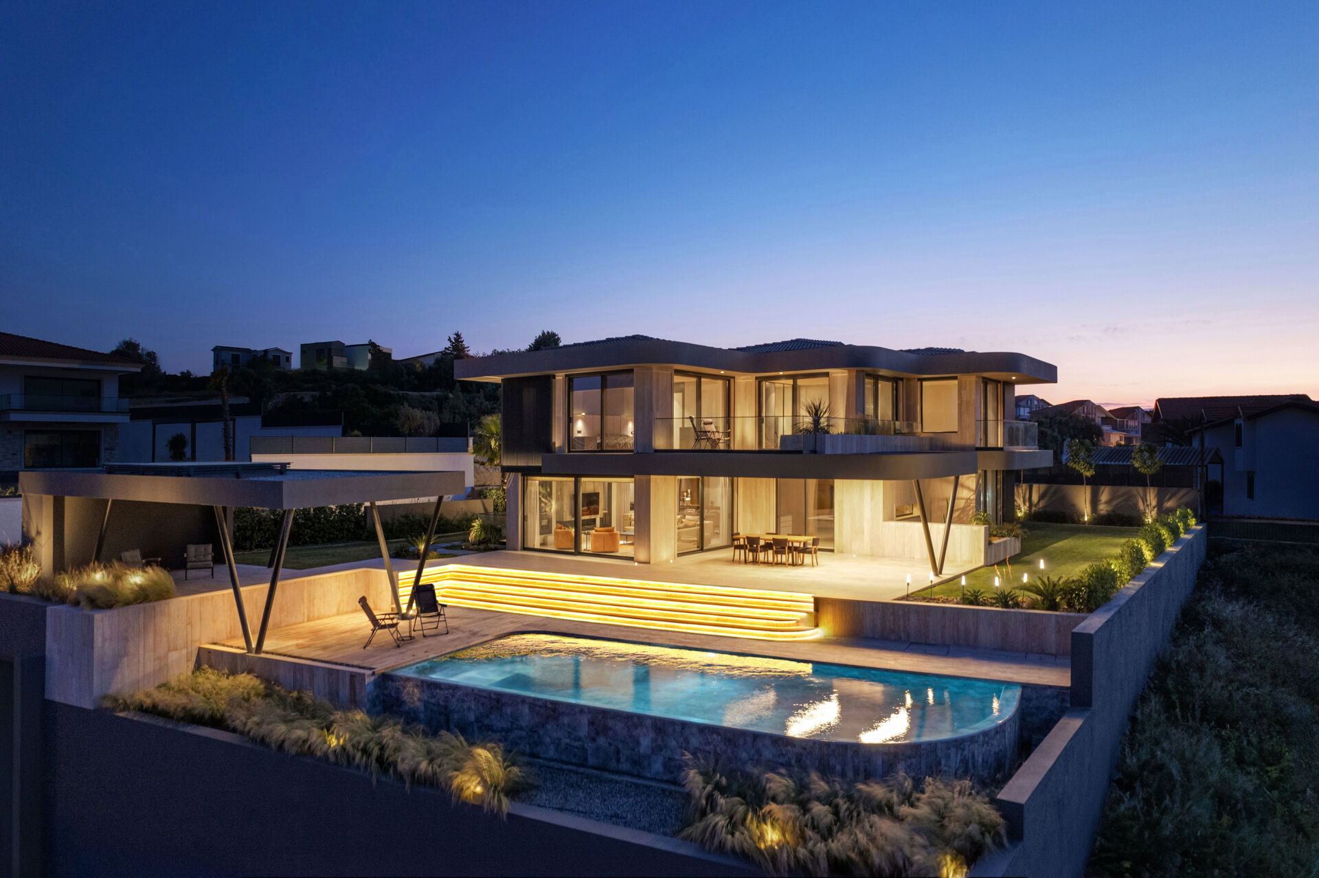 A modern coastal home glowing at night, with soft lighting highlighting sculptural eaves and reflecting across the pool.