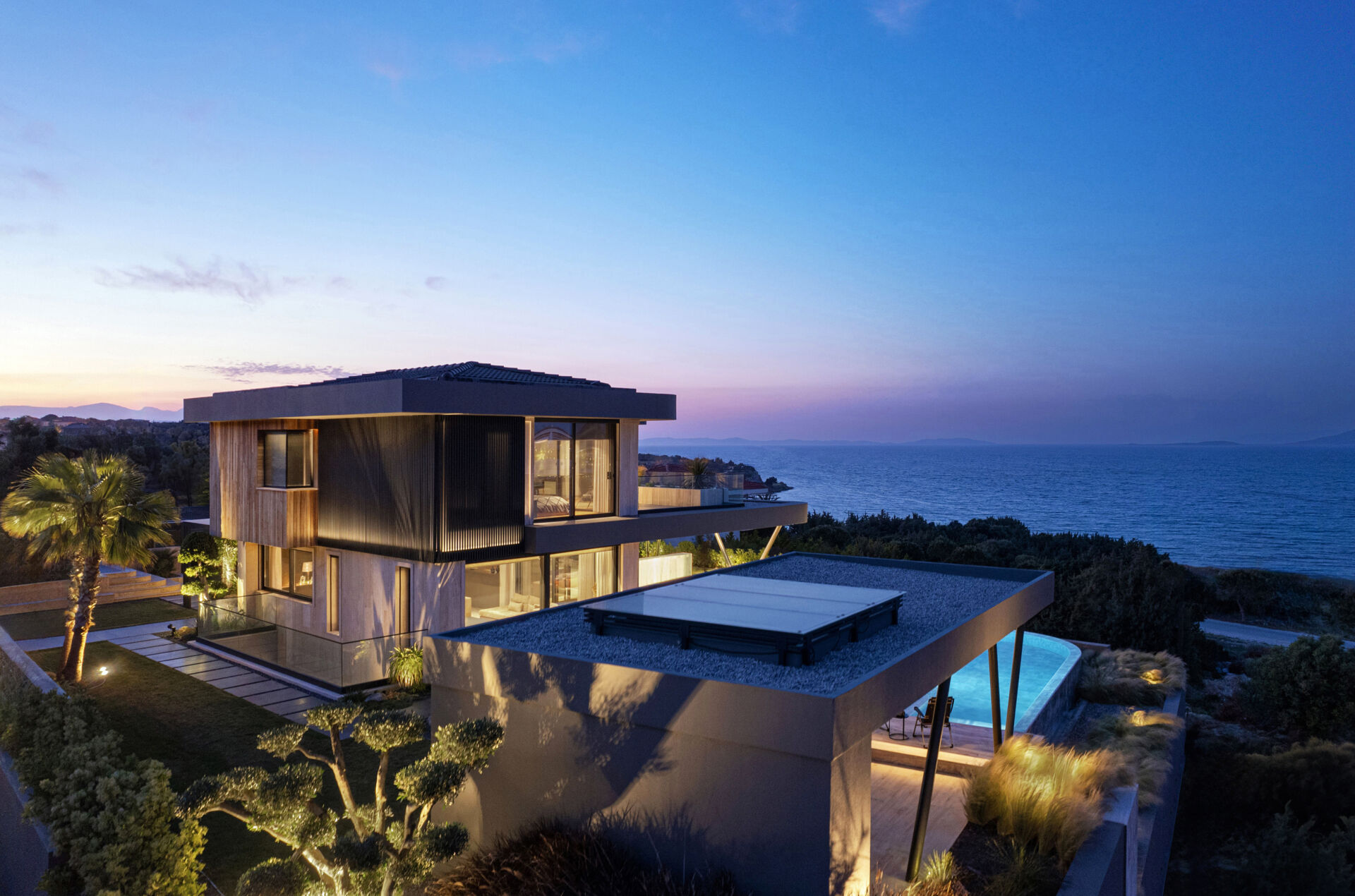 A modern coastal home glowing at night, with soft lighting highlighting sculptural eaves and reflecting across the pool.