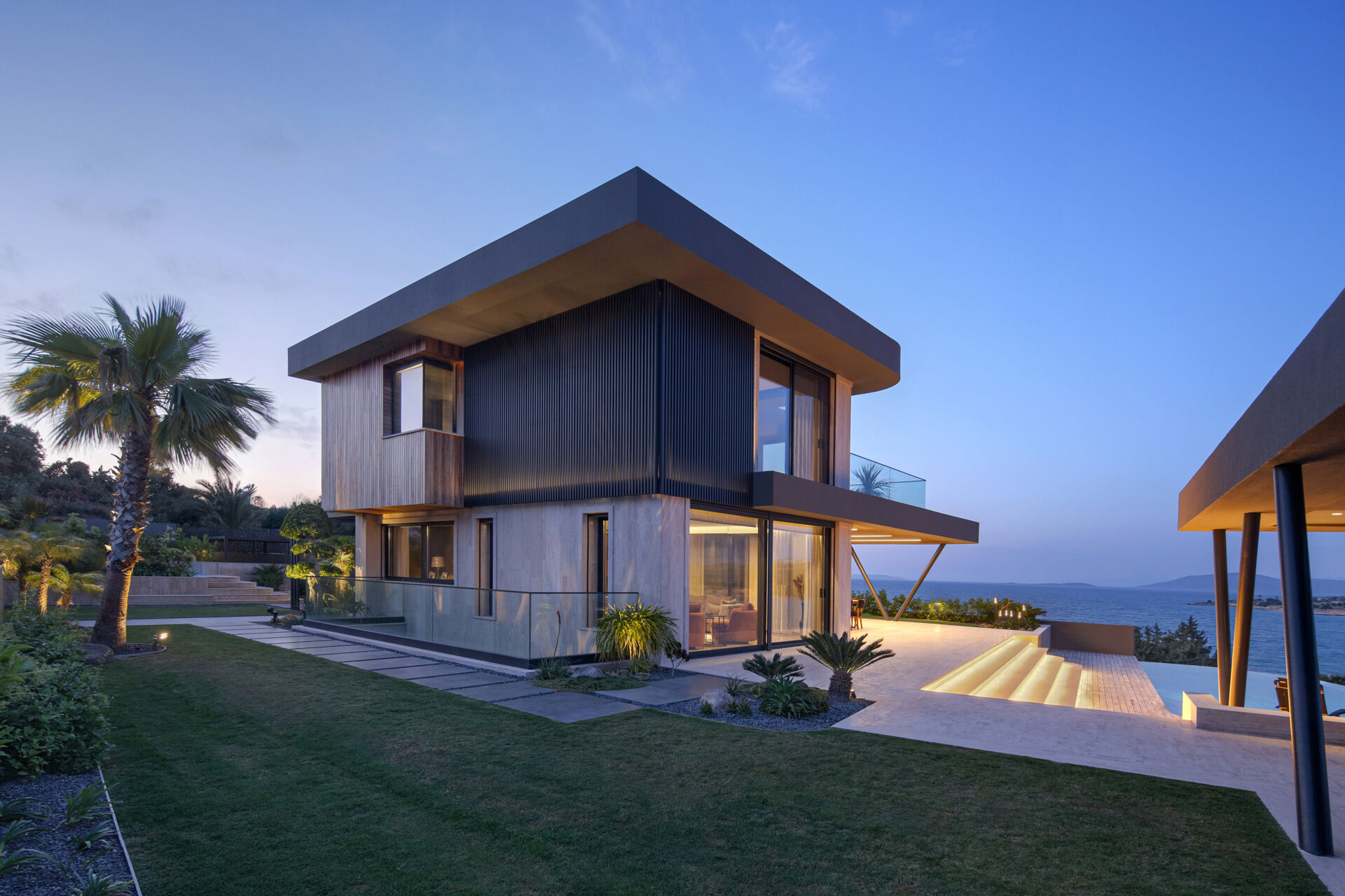 A modern coastal home glowing at night, with soft lighting highlighting sculptural eaves and reflecting across the pool.
