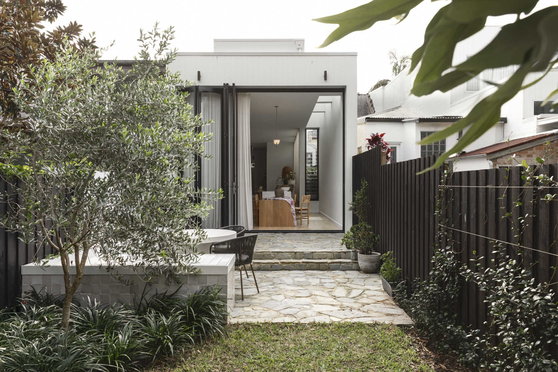 At the rear, the new addition shifts the experience entirely. The architecture becomes more streamlined, opening into a bright, open-plan zone that connects directly to a private terrace.
Stone paving and built-in bench seating define the outdoor space, making it a natural extension of the interior. Large openings blur the boundary between inside and out, allowing the home to expand outward for entertaining or relaxed family time.