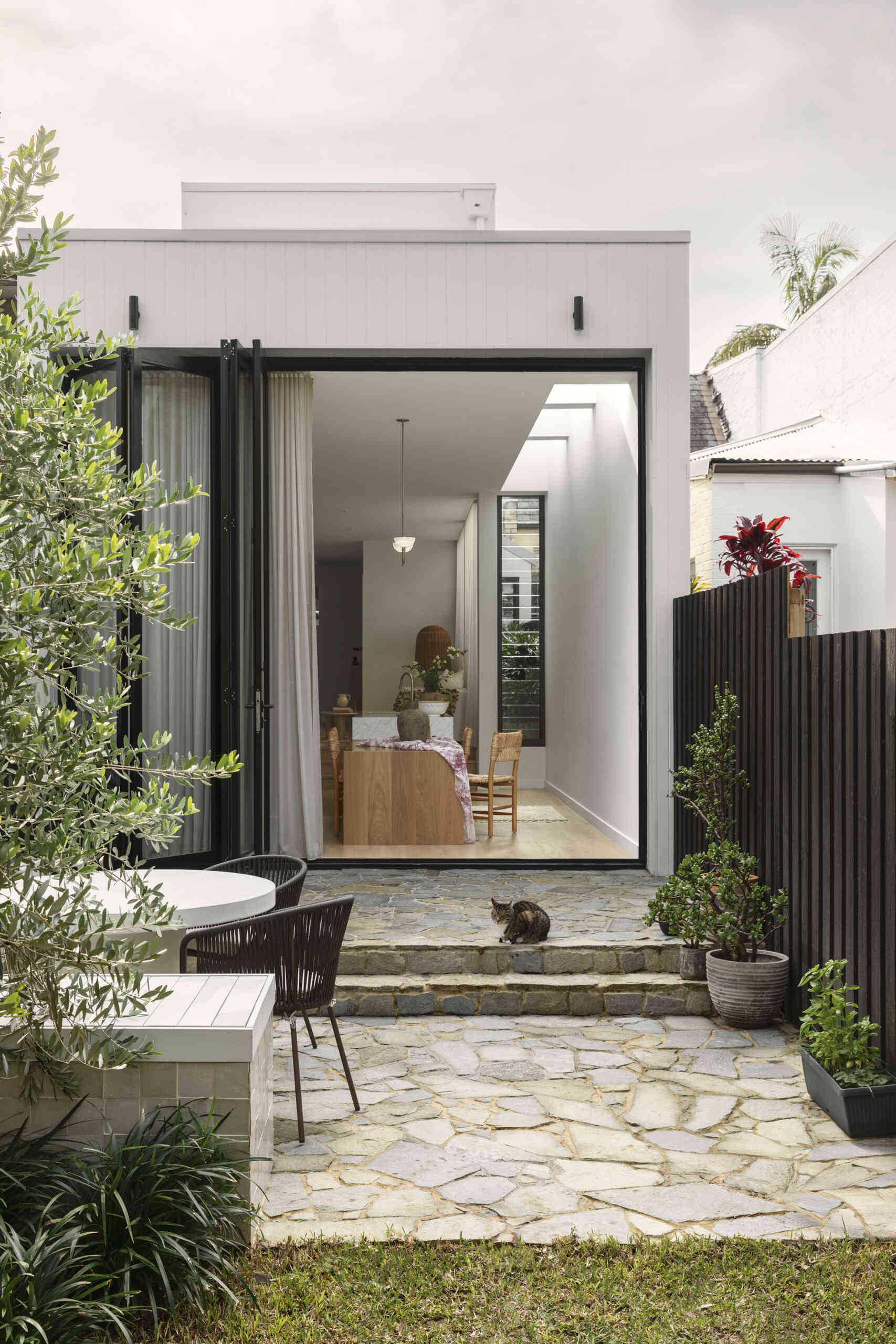At the rear, the new addition shifts the experience entirely. The architecture becomes more streamlined, opening into a bright, open-plan zone that connects directly to a private terrace.
Stone paving and built-in bench seating define the outdoor space, making it a natural extension of the interior. Large openings blur the boundary between inside and out, allowing the home to expand outward for entertaining or relaxed family time.