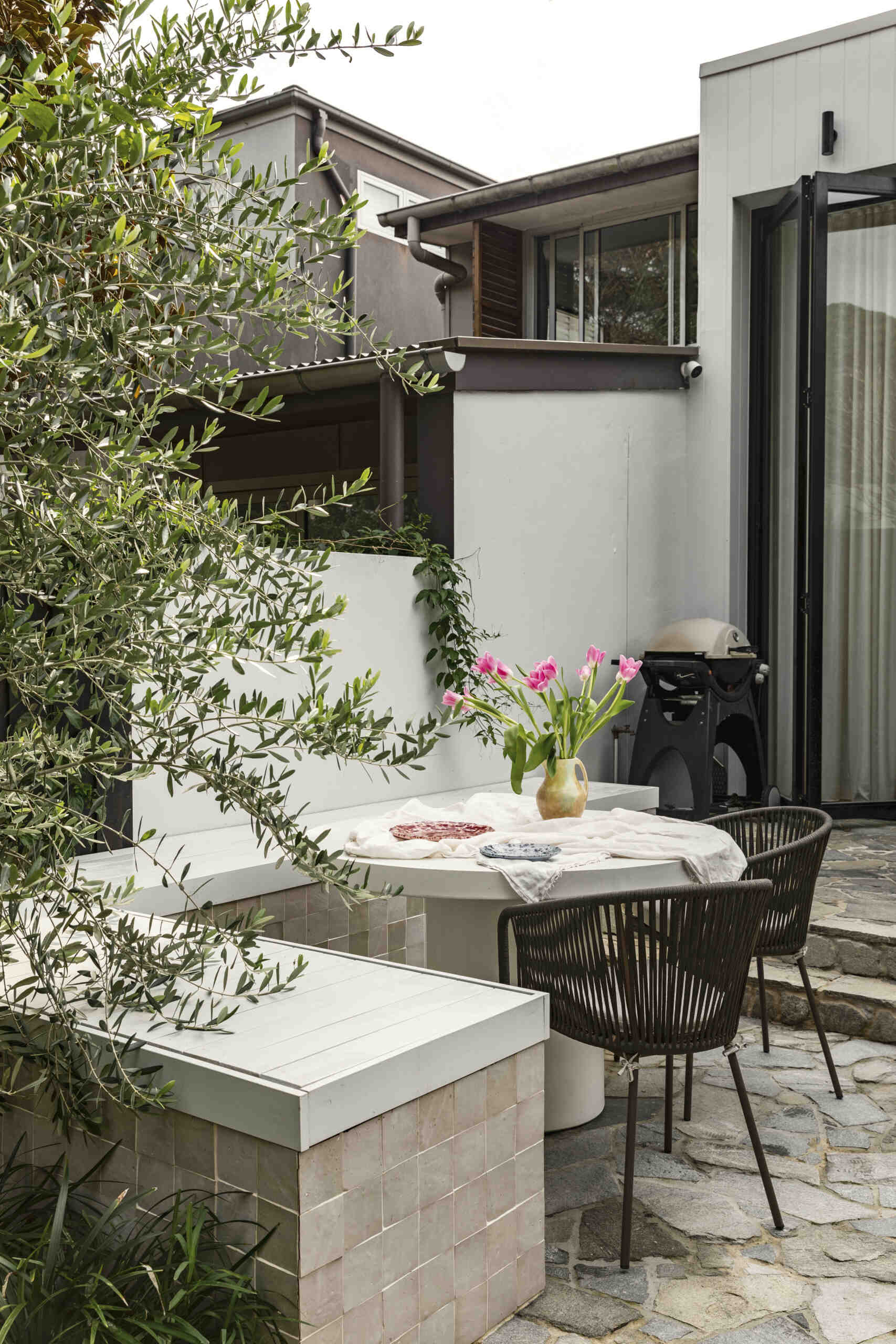 At the rear, the new addition shifts the experience entirely. The architecture becomes more streamlined, opening into a bright, open-plan zone that connects directly to a private terrace.
Stone paving and built-in bench seating define the outdoor space, making it a natural extension of the interior. Large openings blur the boundary between inside and out, allowing the home to expand outward for entertaining or relaxed family time.