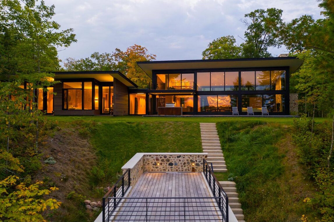 Es una casa de huéspedes moderna de Wisconsin con piedra tosca, amplias paredes de vidrio y vistas panorámicas del lago rodeado por el bosque circundante.