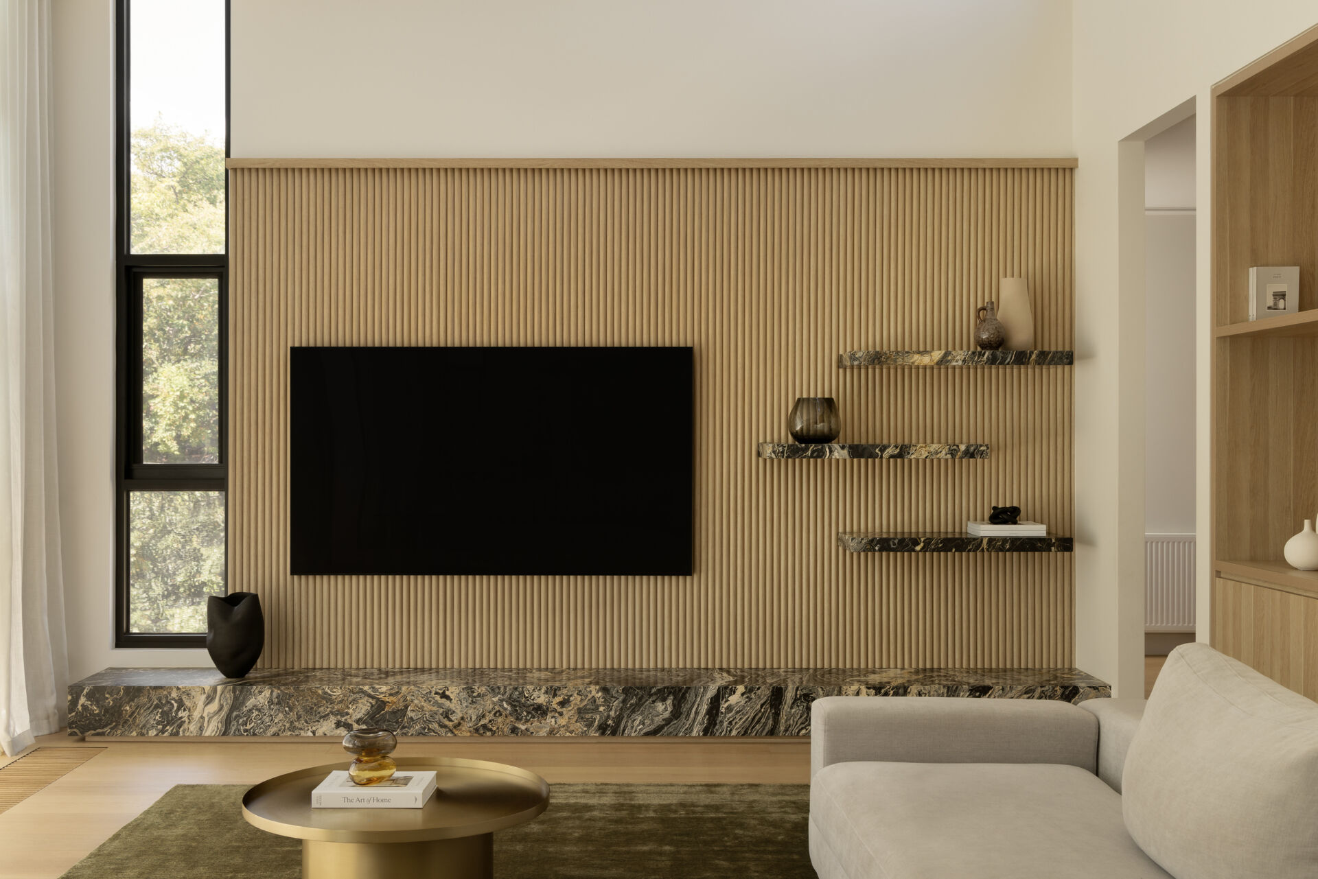 The living room sits beneath a raked ceiling, where high-level windows draw in soft southern light. This creates a balanced atmosphere throughout the day, avoiding harsh contrasts while keeping the space bright. Large openings connect directly to the outdoors, reinforcing the home’s focus on flow and openness.