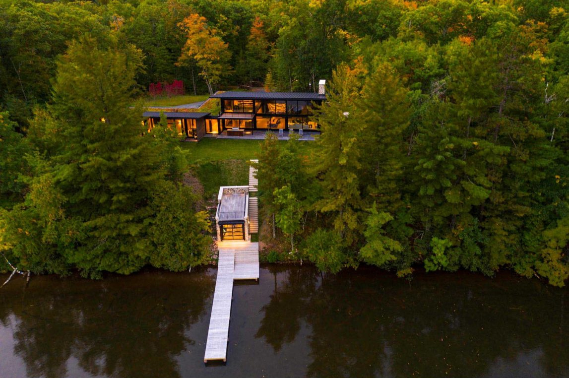Es una casa de huéspedes moderna de Wisconsin con piedra tosca, amplias paredes de vidrio y vistas panorámicas del lago rodeado por el bosque circundante.